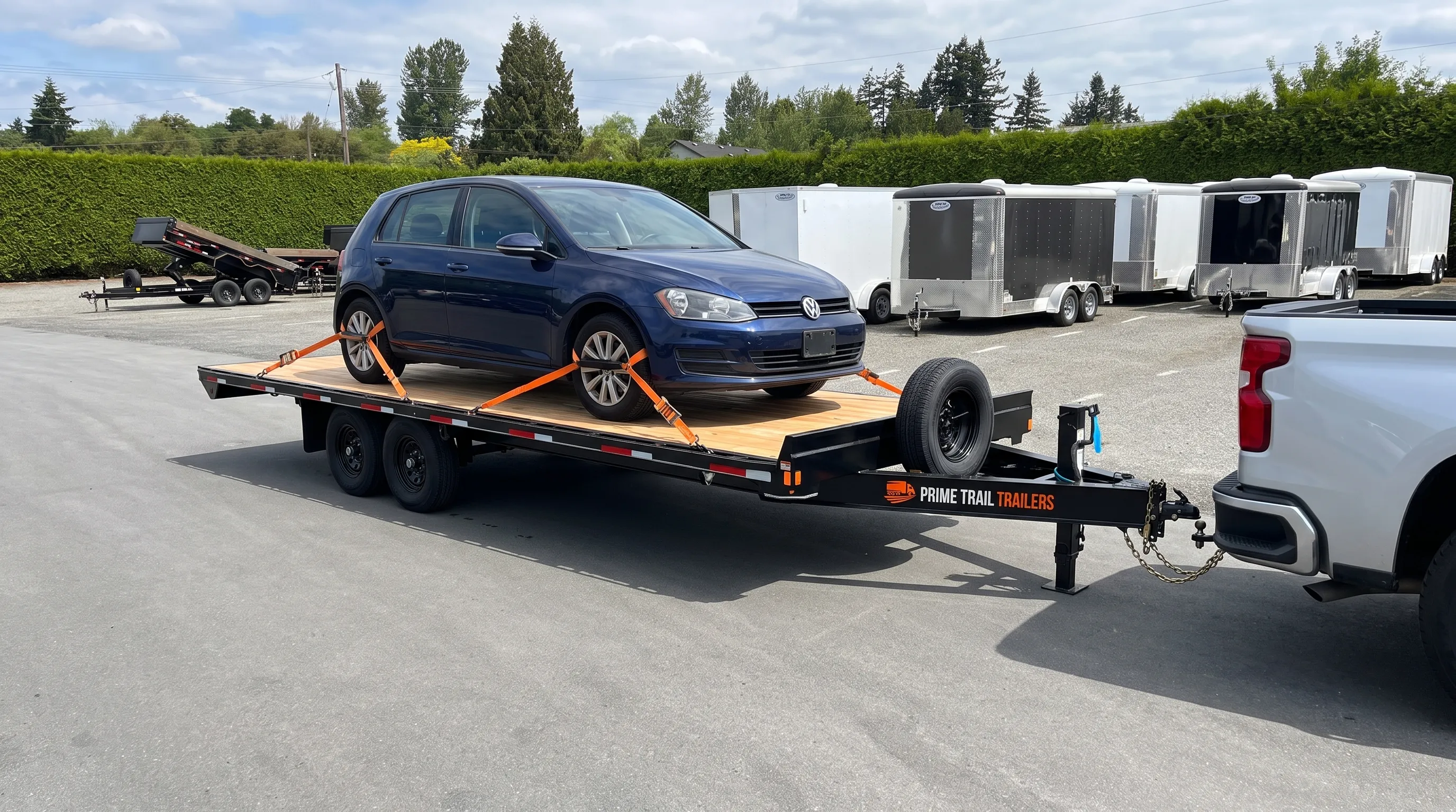 A parked vehicle trailer carries a passenger car with wheel straps secured and a pickup connected at the front.