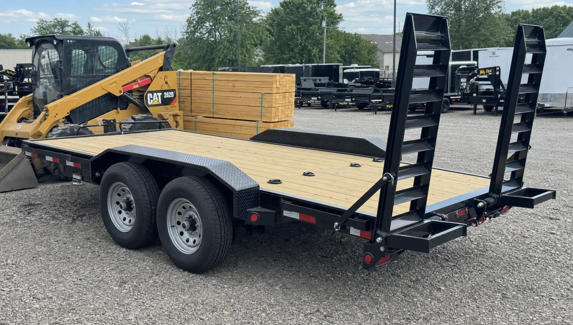 A deck-over trailer on a dealership lot shows its wide open deck, tie-down points and loading ramps beside a skid steer and lumber. A deck-over trailer on a dealership lot shows its wide open deck, tie-down points and loading ramps beside a skid steer and lumber.