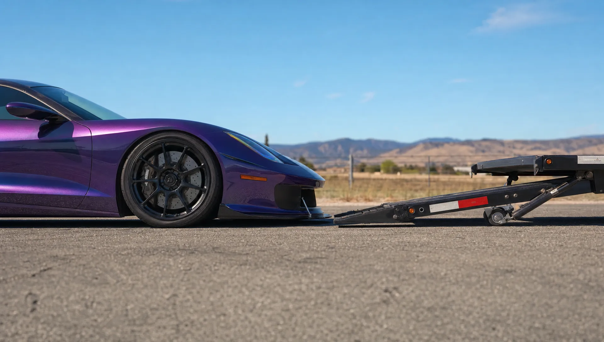 Side view of a low sports car aligned with a hydraulic car trailer, showing the splitter clearance and deck transition on level pavement.