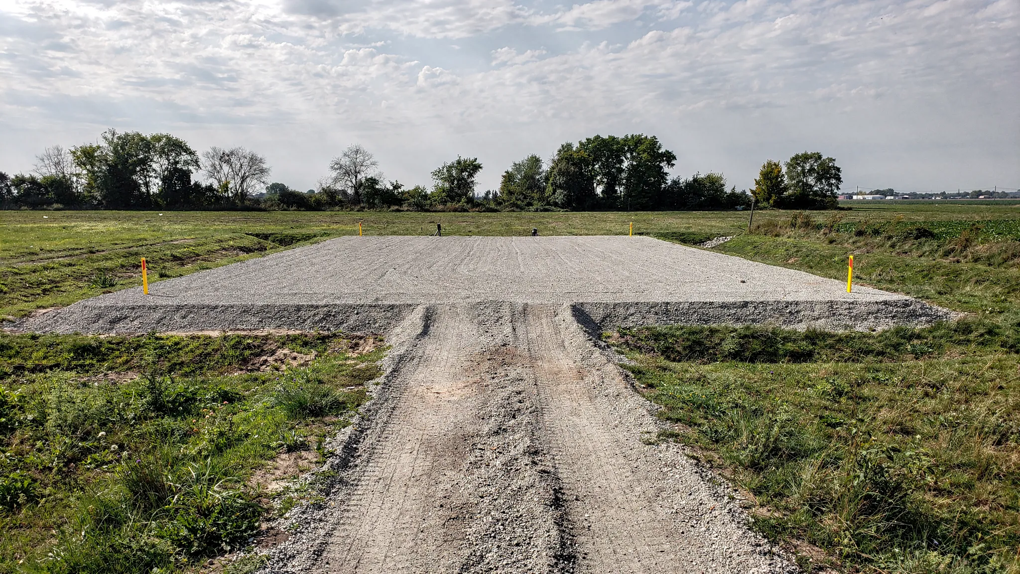 A prepared container drop site with a compacted gravel pad, clear approach path for a delivery truck, and marked corner points where the container will be placed.