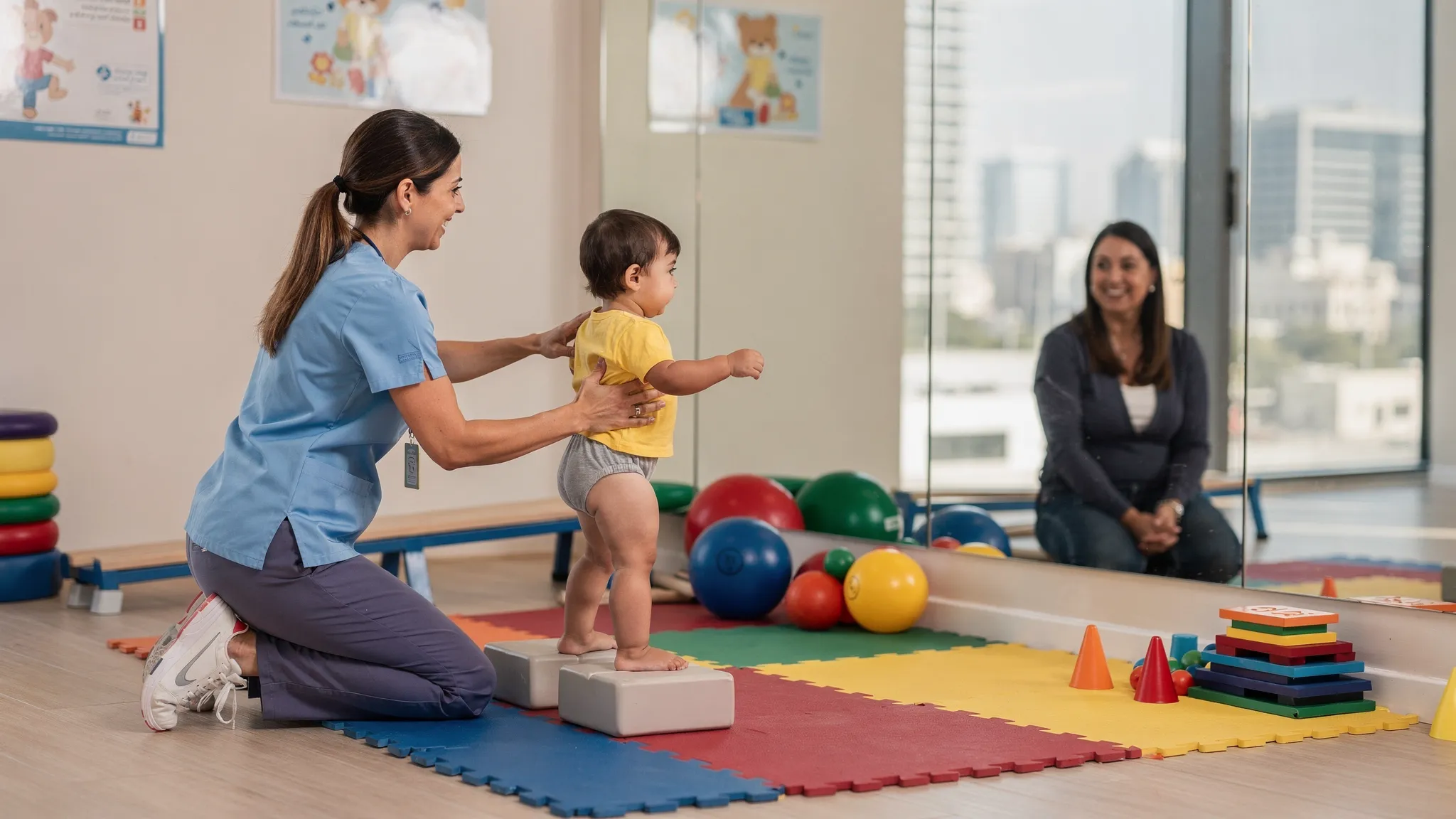 A pediatric physiotherapist in a Dubai clinic helps a toddler practice standing balance using soft blocks and a mirror wall while the parent watches and smiles. Colorful therapy equipment like balance beams, therapy balls and foam mats are visible, creating a warm child-friendly environment.