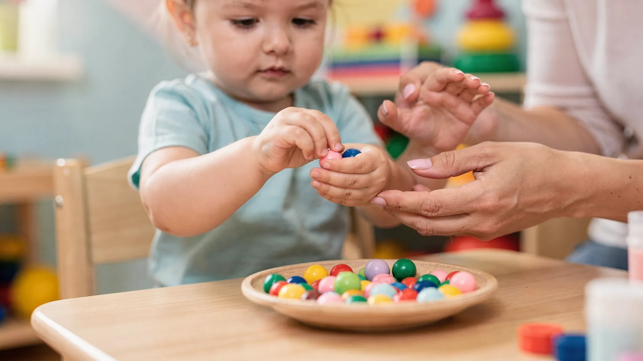 A toddler sits at a child-sized table in a bright therapy room, using a precise pincer grasp to pick up small colorful beads while a parent gently supports the wrist and offers verbal encouragement. Close-up focus on hands and fine motor control.