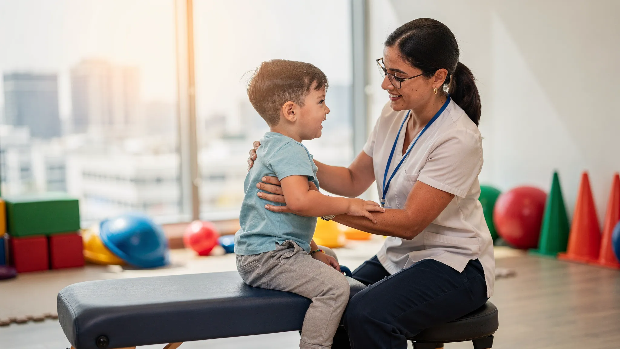 A pediatric physiotherapist guides a preschool child through a Dynamic Movement Intervention session on a low bench, using light distal holds at the pelvis and legs to elicit protective responses and midline control, with colorful therapy equipment visible in a bright clinic room in Dubai.