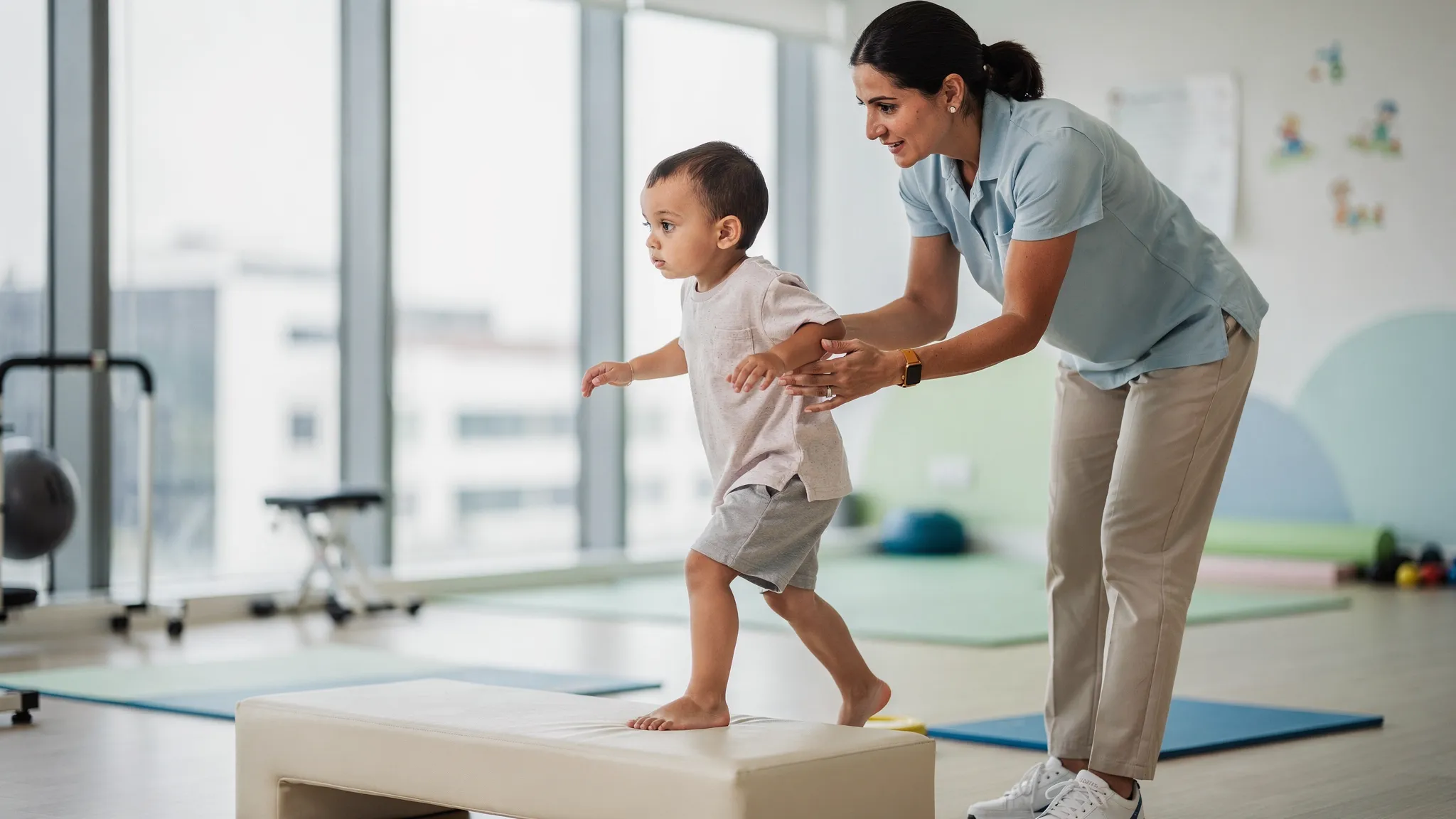A toddler practices cuevas medek exercise with a physiotherapist who provides a light distal hold at the ankles while the child steps forward off a foam block, capturing a protective step response and upright alignment in a bright Dubai clinic.