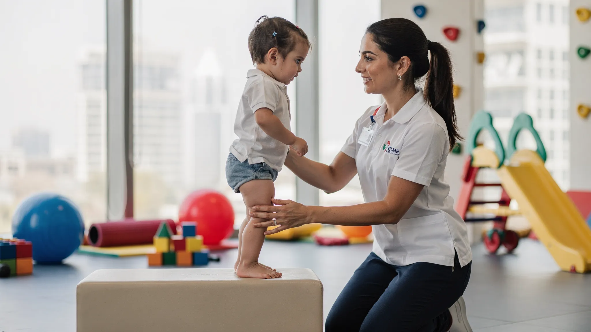 A pediatric physiotherapist in Dubai guiding a young child with cerebral palsy through a CME exercise using distal support at the ankles while the child stands on a foam block, with bright therapy equipment in the background.
