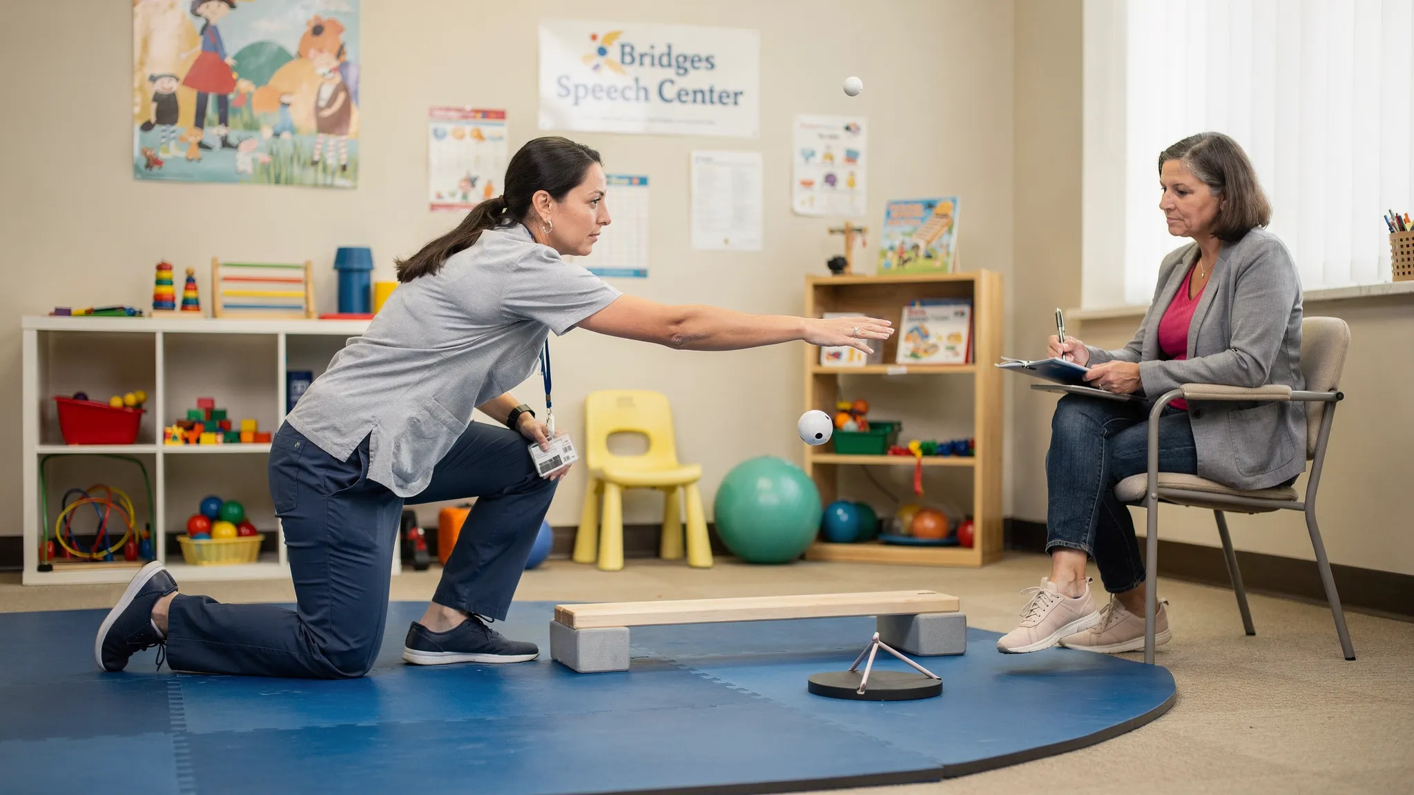 Parent coaching during CME therapy at Bridges Speech Center, with a therapist demonstrating a balance reach task on a soft mat while the parent observes and takes notes in a child-friendly rehab room.