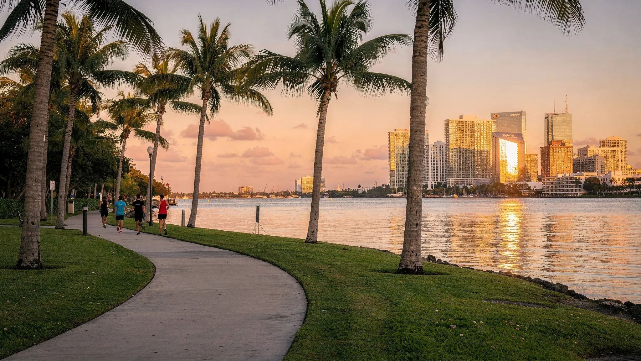 A scenic walking path on Belle Isle beside Biscayne Bay at sunrise, with palm trees, a few joggers in the distance, and the Miami skyline glowing across the water.