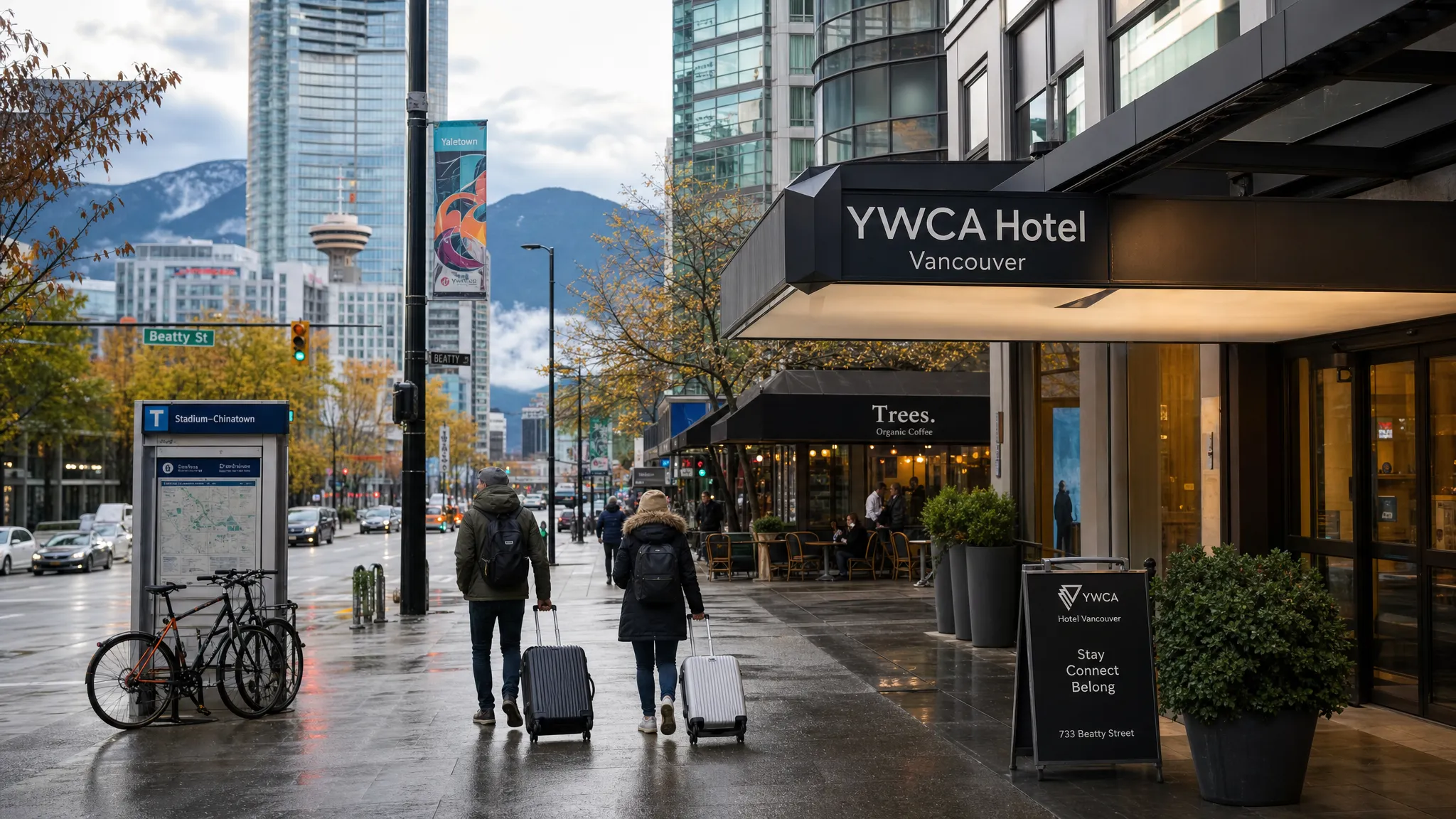 A rainy Vancouver street scene near a compact downtown hotel, with travelers walking past cafes, glass towers, trees, and distant mountain silhouettes, showing how central hotel location connects transit, food, and sightseeing.