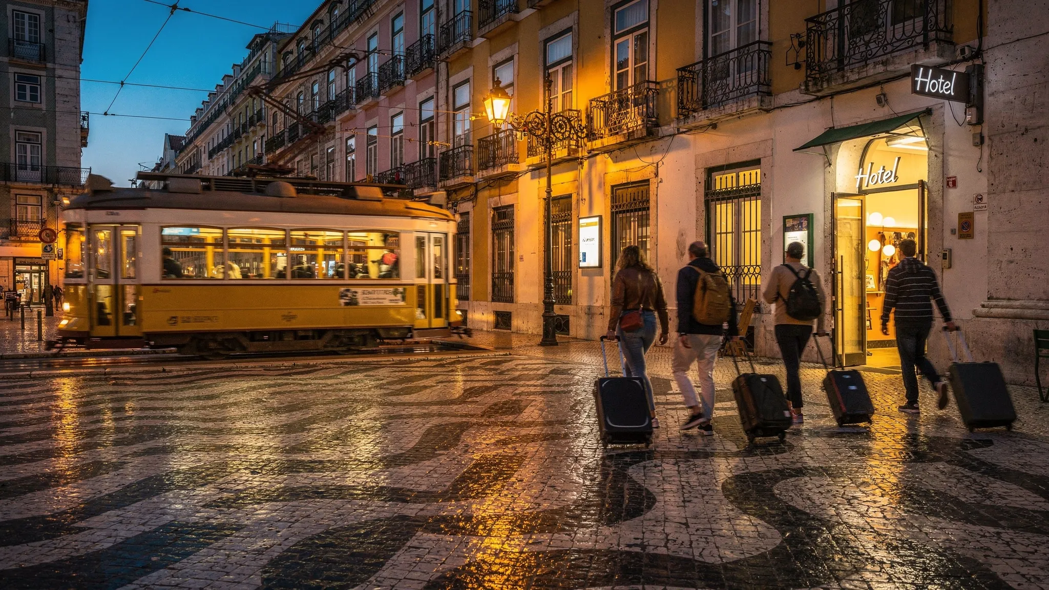 Evening in Lisbon near Rossio and Baixa, with patterned pavement, warm streetlights reflecting on historic facades, a classic yellow tram passing in the background, and travelers walking with small suitcases toward a softly lit hotel entrance.