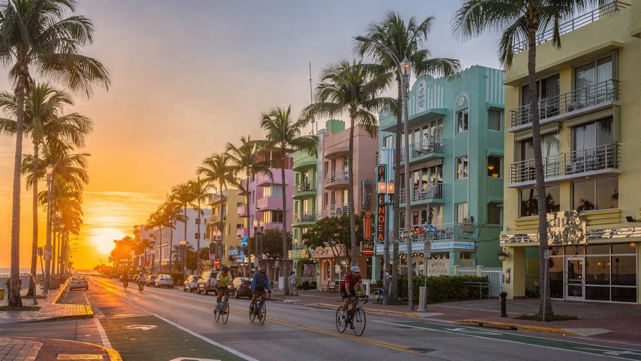 Sunrise on Ocean Drive in Miami Beach with pastel Art Deco hotels, palm trees lining the street, soft orange light reflecting on windows, and a few early cyclists passing by.