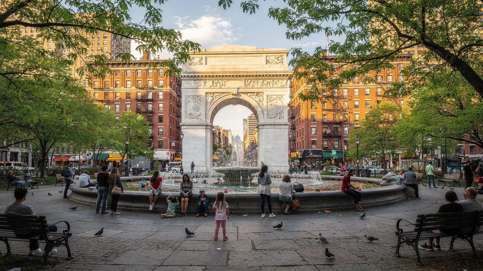 Washington Square Park on a mild afternoon with the arch in the background, people gathered around the fountain, leafy trees framing the scene, and warm light reflecting off nearby brick buildings.