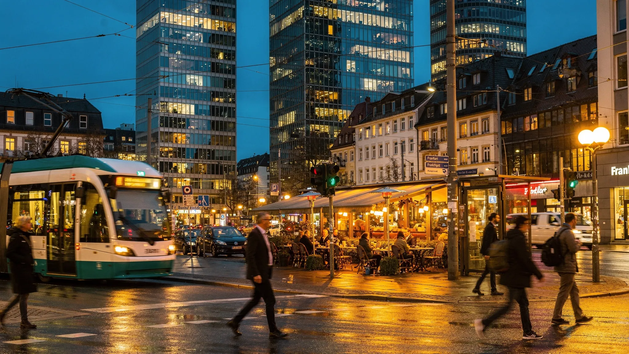 An evening street scene in central Frankfurt with modern glass towers in the distance, warm lights from sidewalk cafes, and a tram passing through a busy intersection.