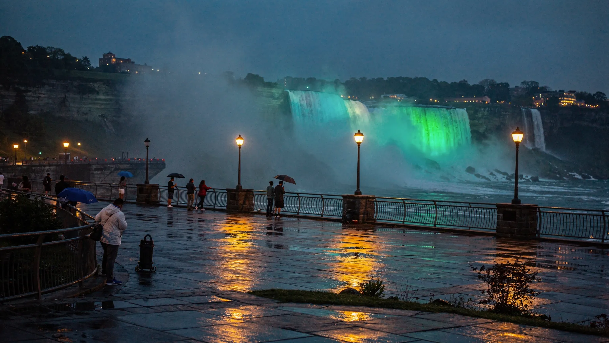 A misty weeknight scene at Niagara Falls State Park at dusk, with fewer people on the overlook railings, wet stone walkways reflecting soft streetlights, and the illuminated falls in the background.