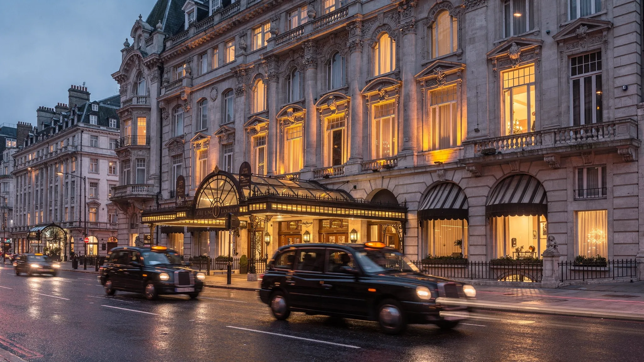 An evening view of The Langham’s grand historic facade on Portland Place in London, with warm golden light glowing through tall windows, a classic entrance canopy, and a few black taxis passing by on a quiet, elegant street.