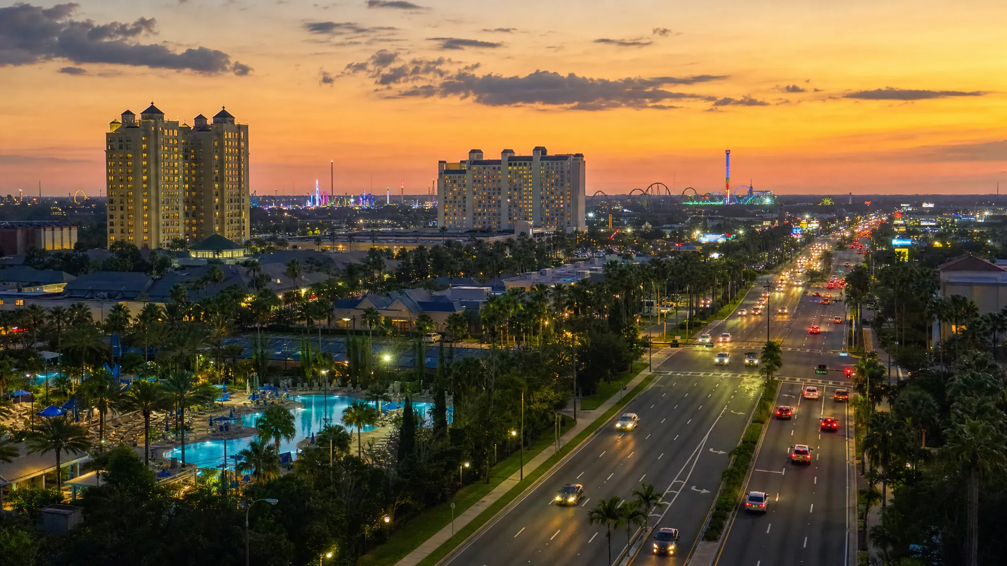 Wide landscape view of Orlando at sunset from a slightly elevated roadside angle, with resort pools, palm-lined roads, distant theme park lights, hotel towers, and cars moving along a broad boulevard to show how location and transportation shape the stay.