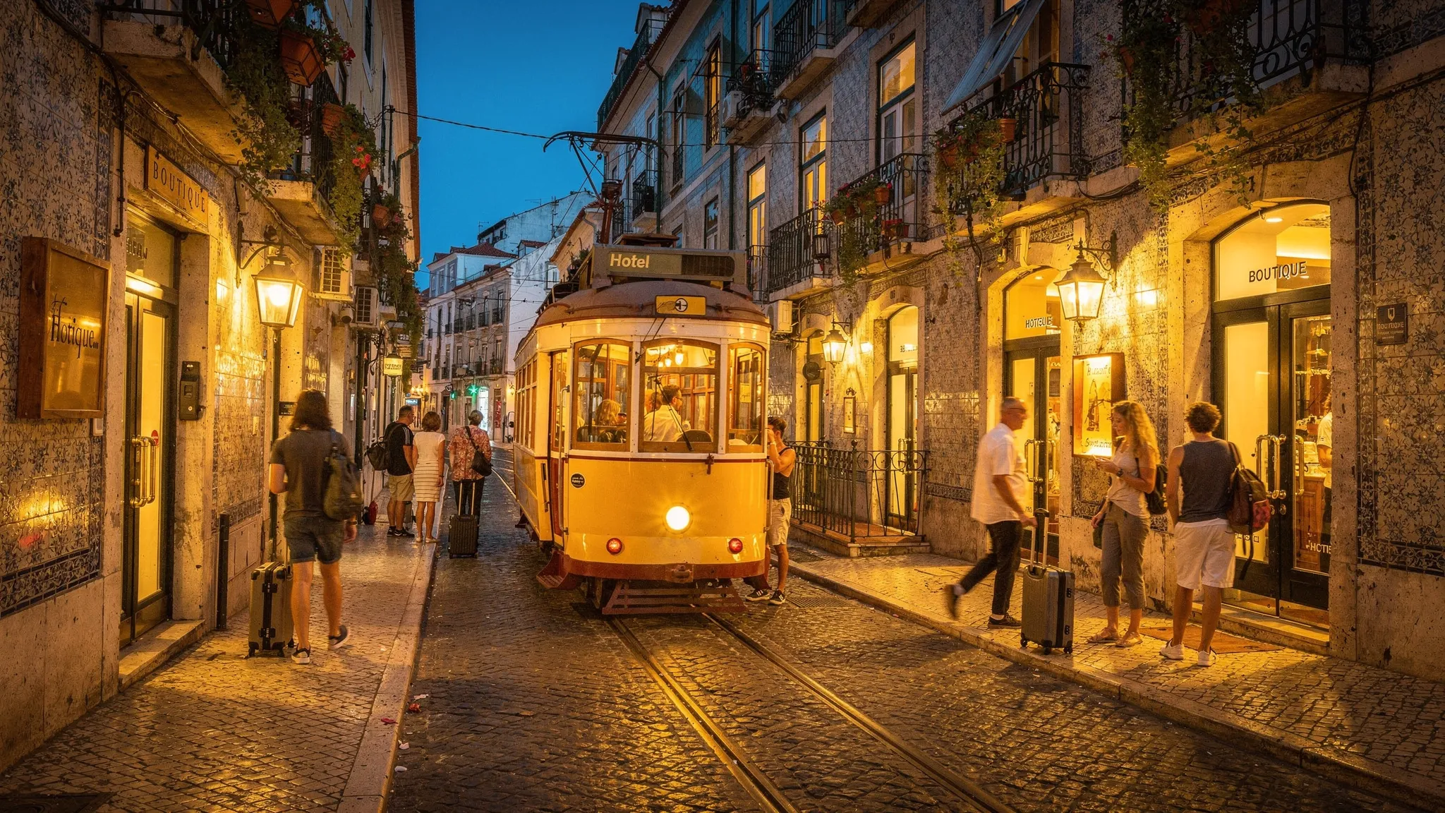 A narrow Lisbon street at dusk with tiled buildings, a yellow tram, travelers with small suitcases, and boutique hotel entrances glowing under warm lights.