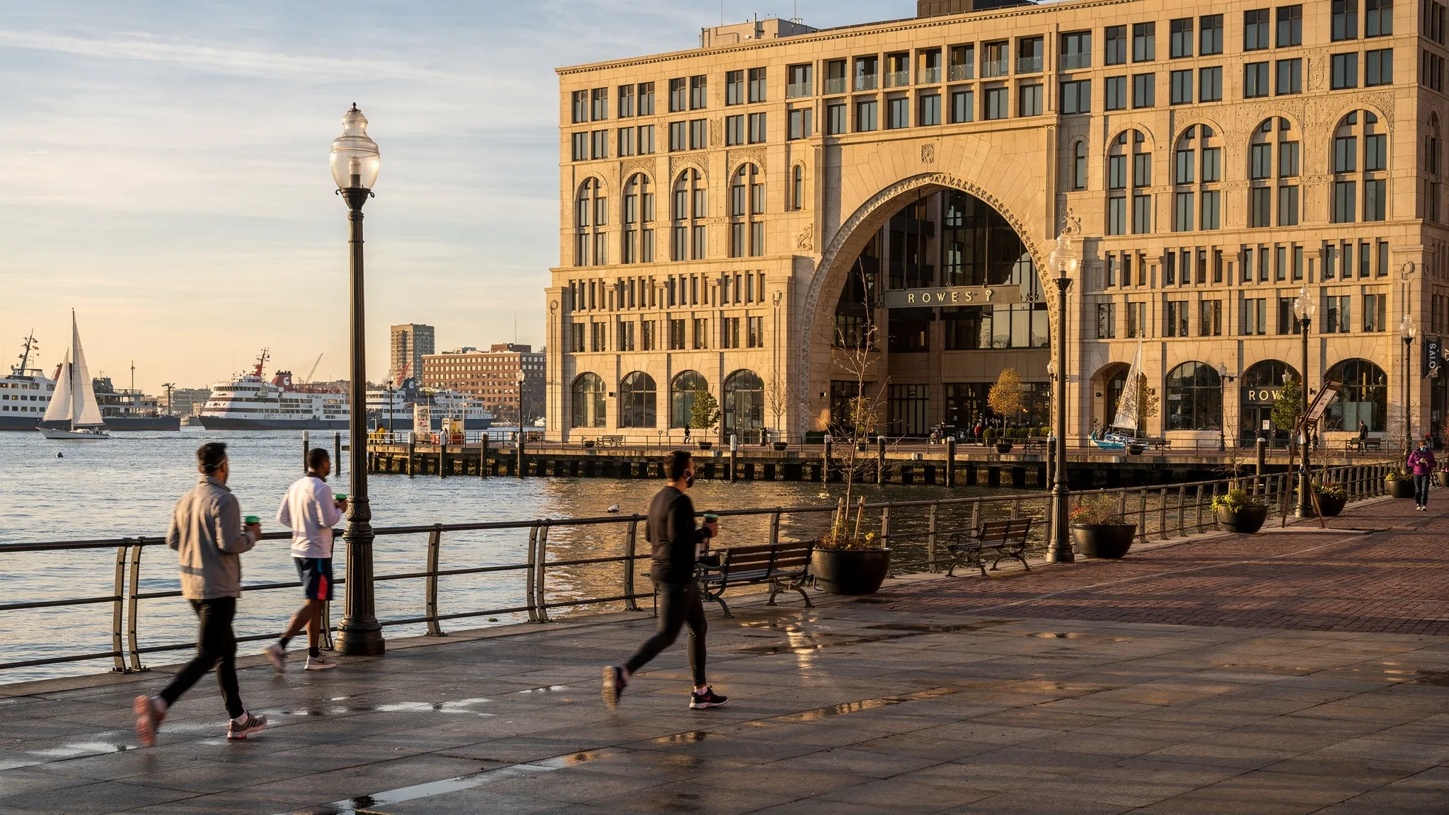 A crisp morning scene at Boston’s Rowes Wharf with the waterfront promenade in the foreground, walkers and joggers passing by, the iconic archway entrance of a harborfront hotel in the midground, and ferries and sailboats on the water under soft golden light.