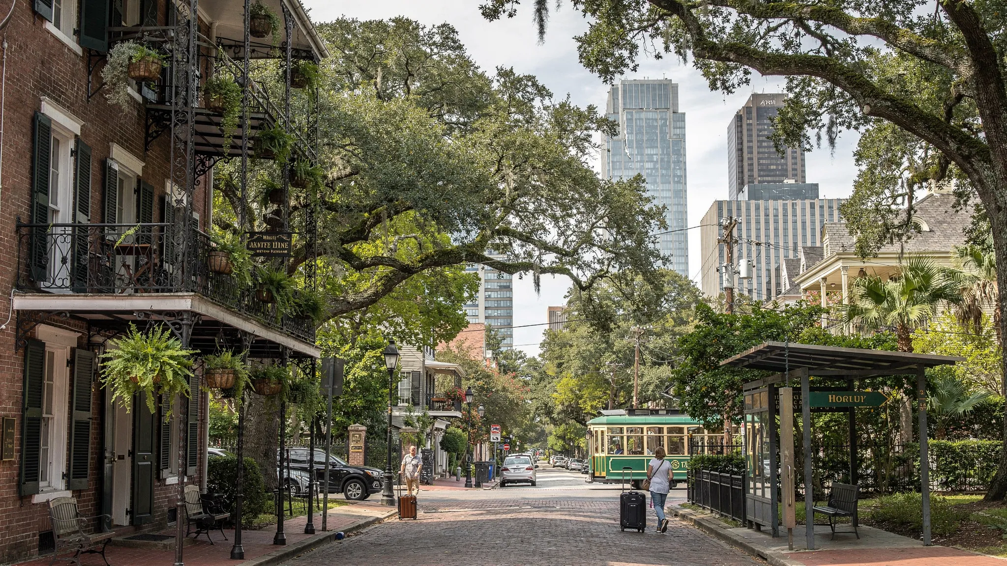 Street-level view of New Orleans hotel neighborhoods, with a French Quarter balcony hotel, a quiet Garden District streetcar stop, and modern CBD hotel towers in the distance.