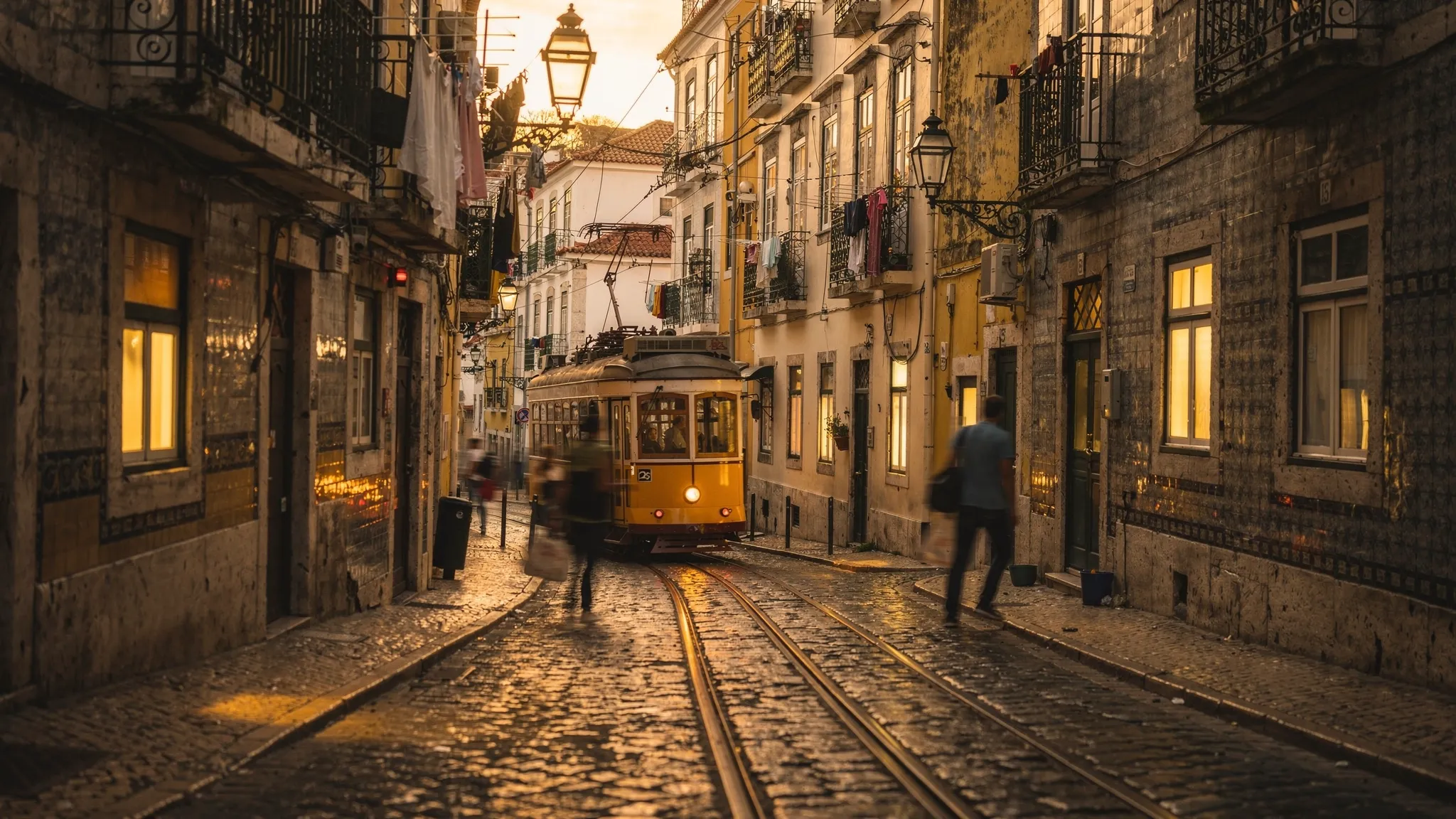 A street-level scene in Lisbon’s Alfama at golden hour with narrow cobblestone lanes, tiled building facades, warm window light, and a classic yellow tram passing in the background. The mood feels intimate and atmospheric, emphasizing neighborhood texture.