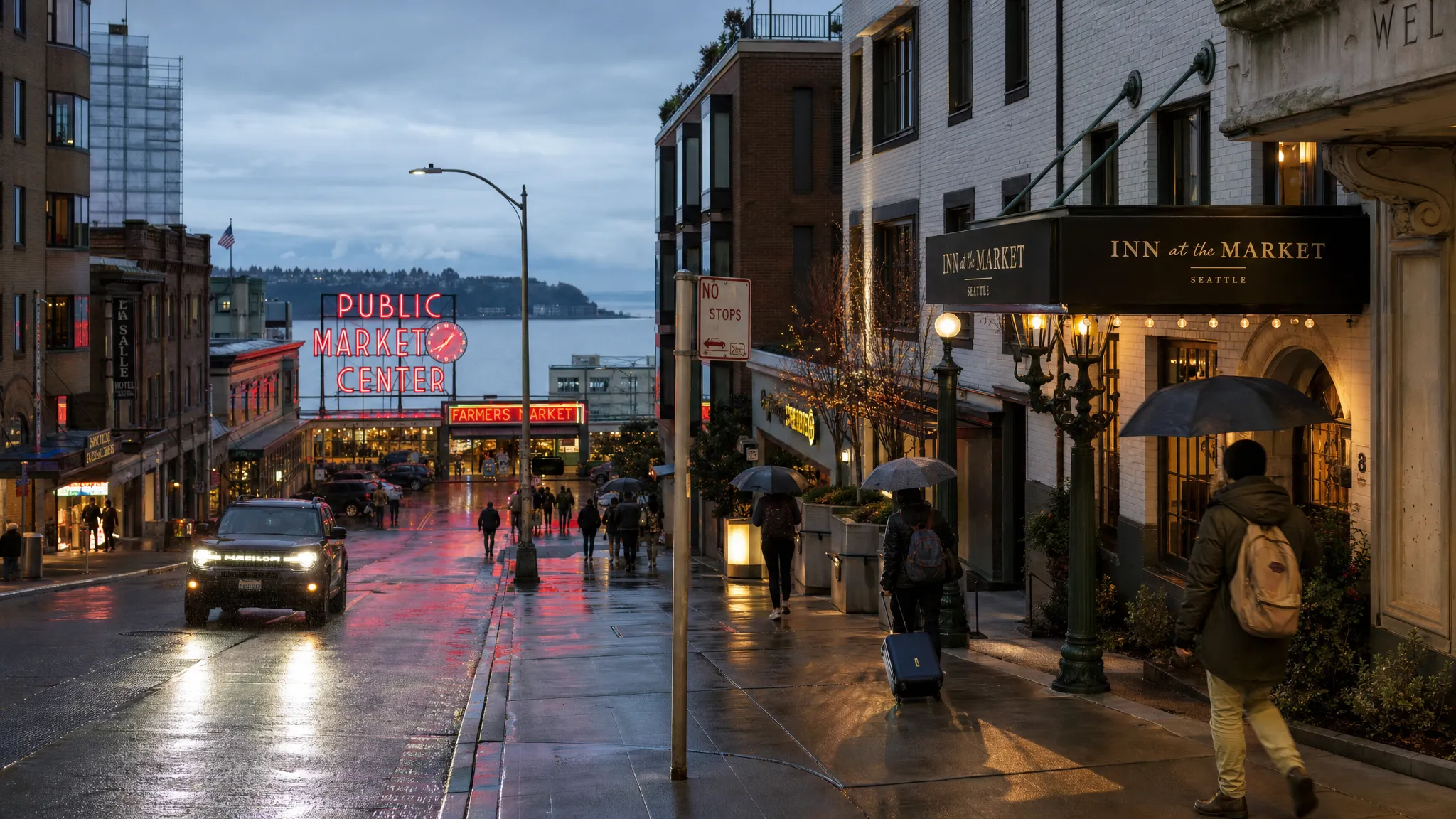 A rainy Seattle street at dusk with hotel entrances, market lights, travelers carrying umbrellas, and the waterfront faintly visible beyond the buildings.