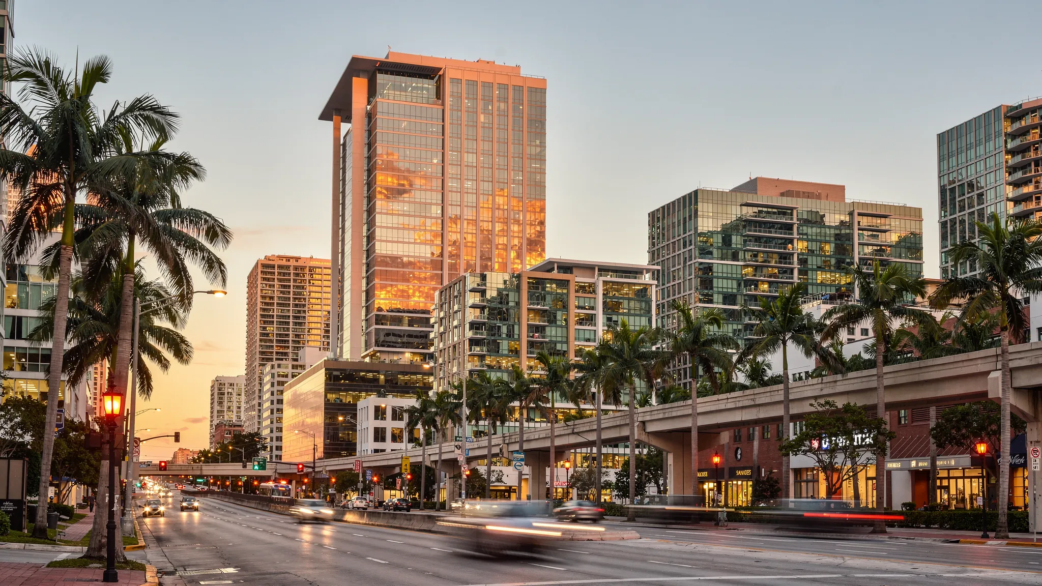 A modern high-rise hotel tower rises above Brickell in Miami at golden hour, surrounded by glass buildings, palm trees, elevated transit tracks, and warm city lights beginning to glow.