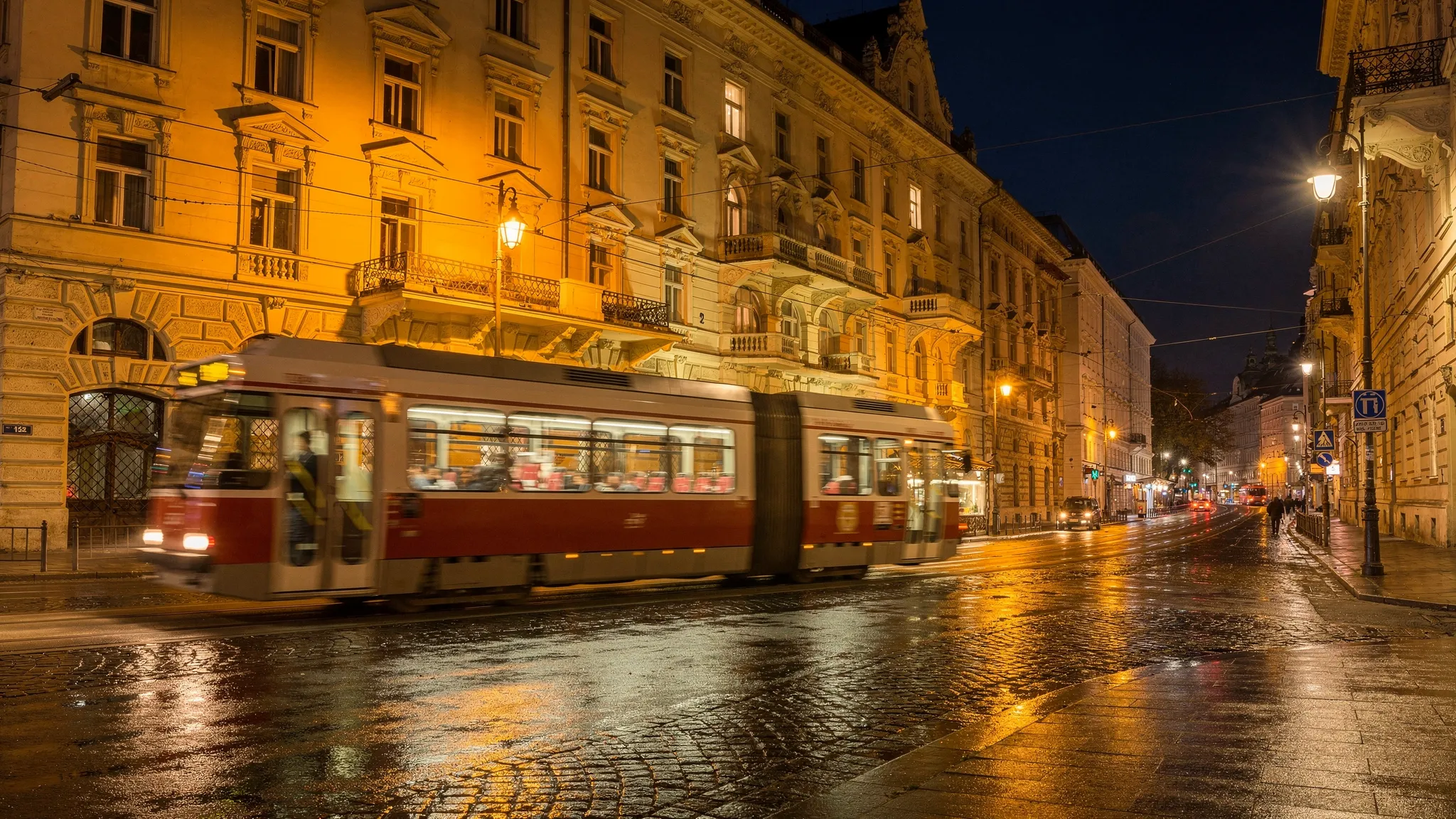 A nighttime street scene in Vienna near the Ringstraße with softly lit historic facades, a passing red-and-white tram, wet cobblestones reflecting warm streetlights, and a quiet side street turning away from the main road.