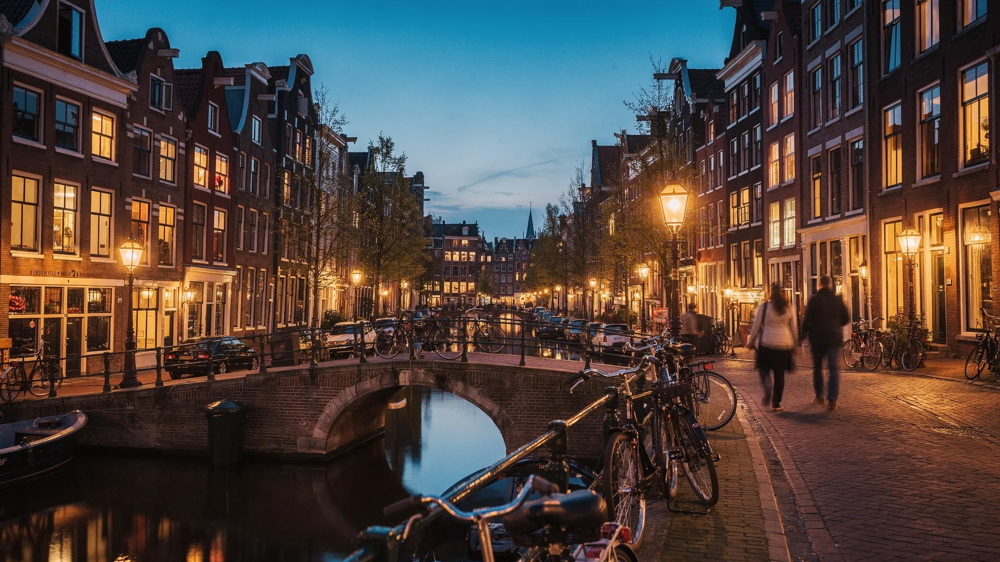Evening in Amsterdam’s canal belt with warm lights reflecting on the water, narrow historic buildings, a small bridge, and a quiet street scene suggesting a calm post-meeting walk.