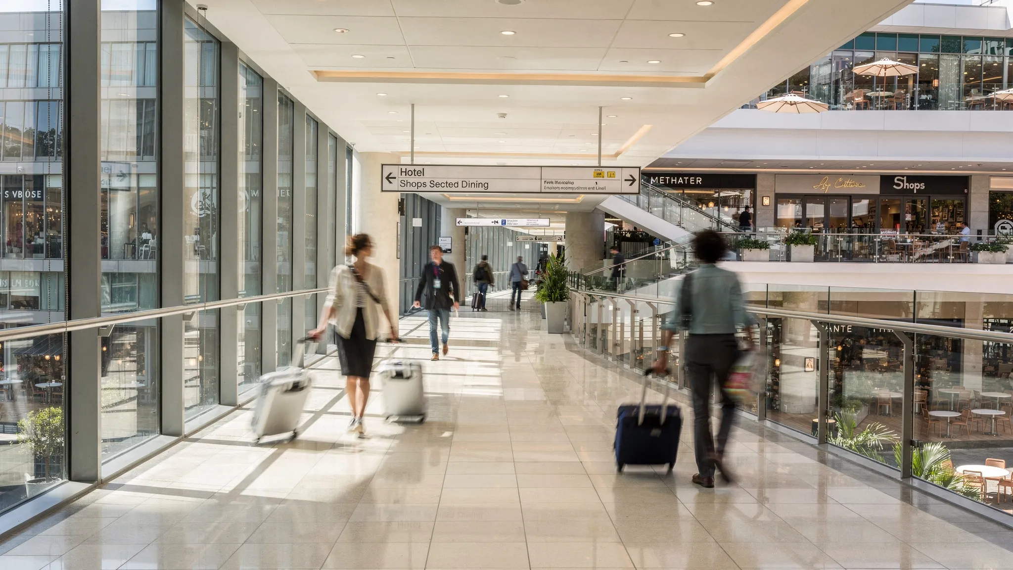 An enclosed skyway corridor connecting a modern hotel to a large shopping and dining complex, with bright lighting, clean signage, and travelers walking with small suitcases.