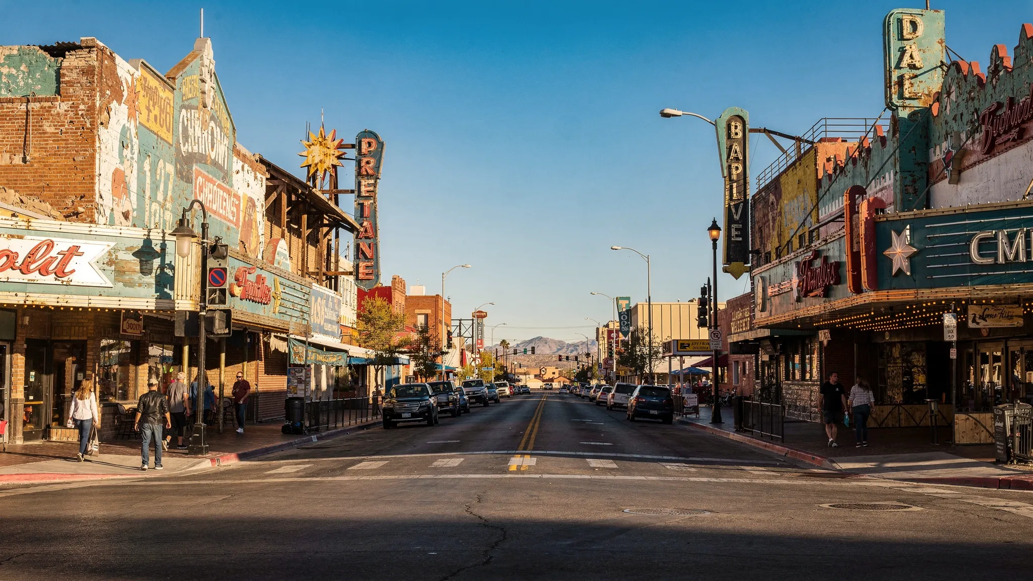 A bright Sunday morning scene in Fremont East with vintage neon signs, low-rise casino facades, a mostly empty street with a few pedestrians, and desert-blue sky highlighting old brick and metal details.