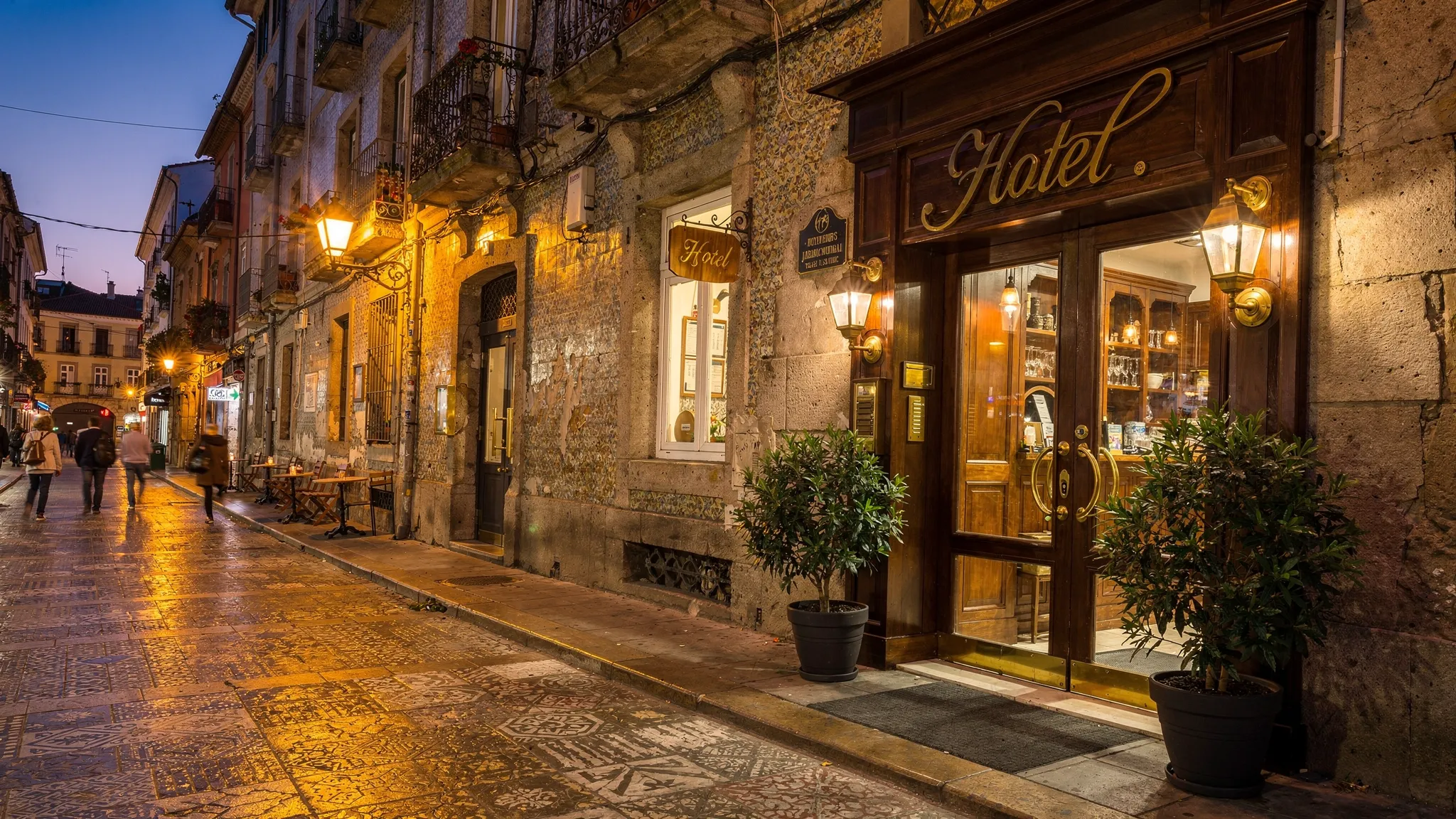 A dusk street scene in a historic European neighborhood with patterned stone sidewalks, tiled building facades, warm café lighting, and a small boutique hotel entrance with brass signage and potted plants.