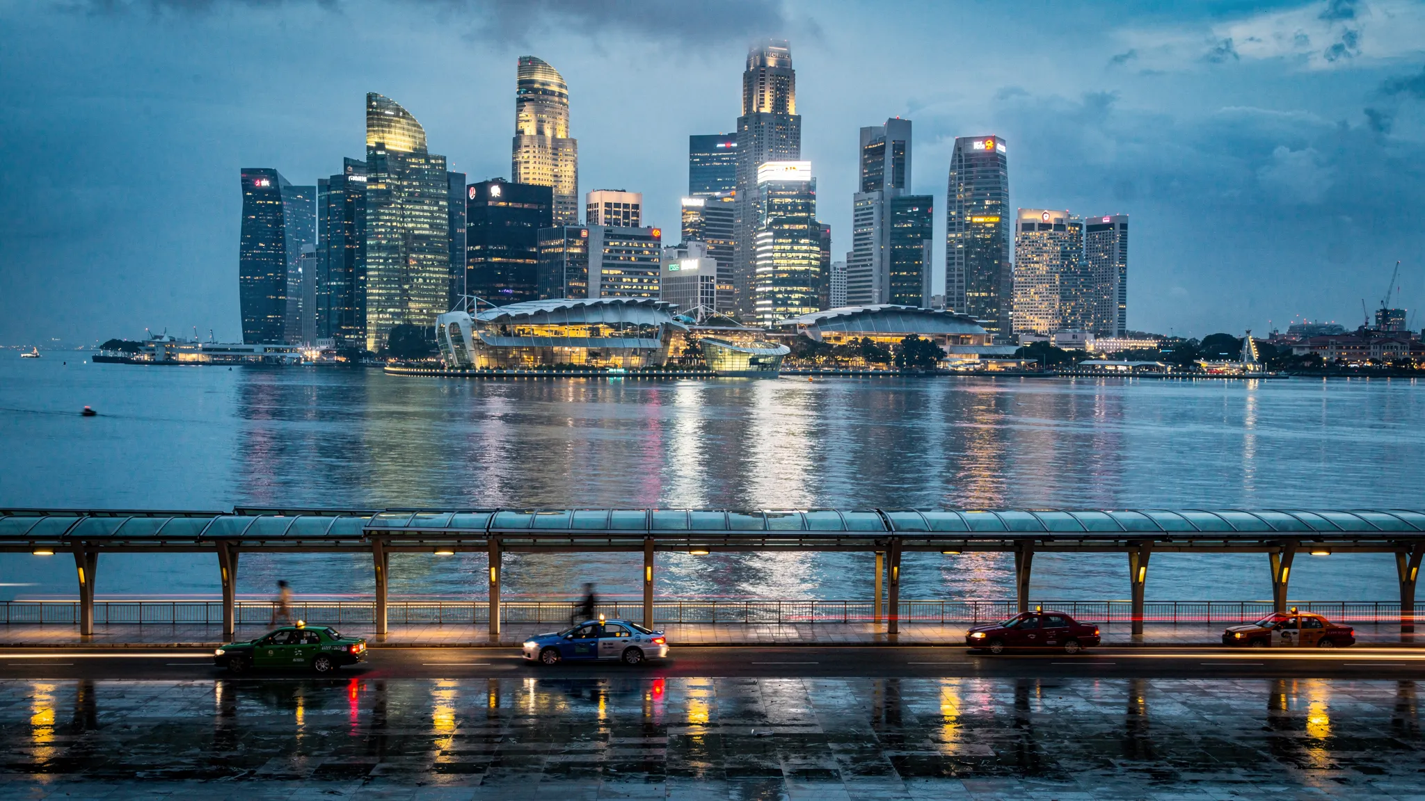 A rainy blue-hour view of Singapore’s Marina Bay skyline with office towers and hotel lights reflecting on calm water, with a covered promenade in the foreground and a few taxis passing by.