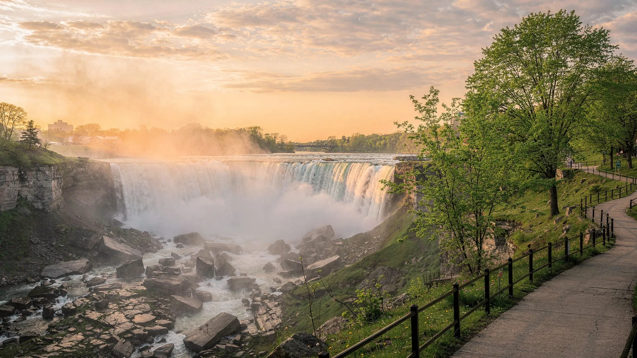 A misty sunrise view of Niagara Falls from the New York side, with the rocky gorge in the foreground, soft golden light on the water, and a calm walking path along the park edge.