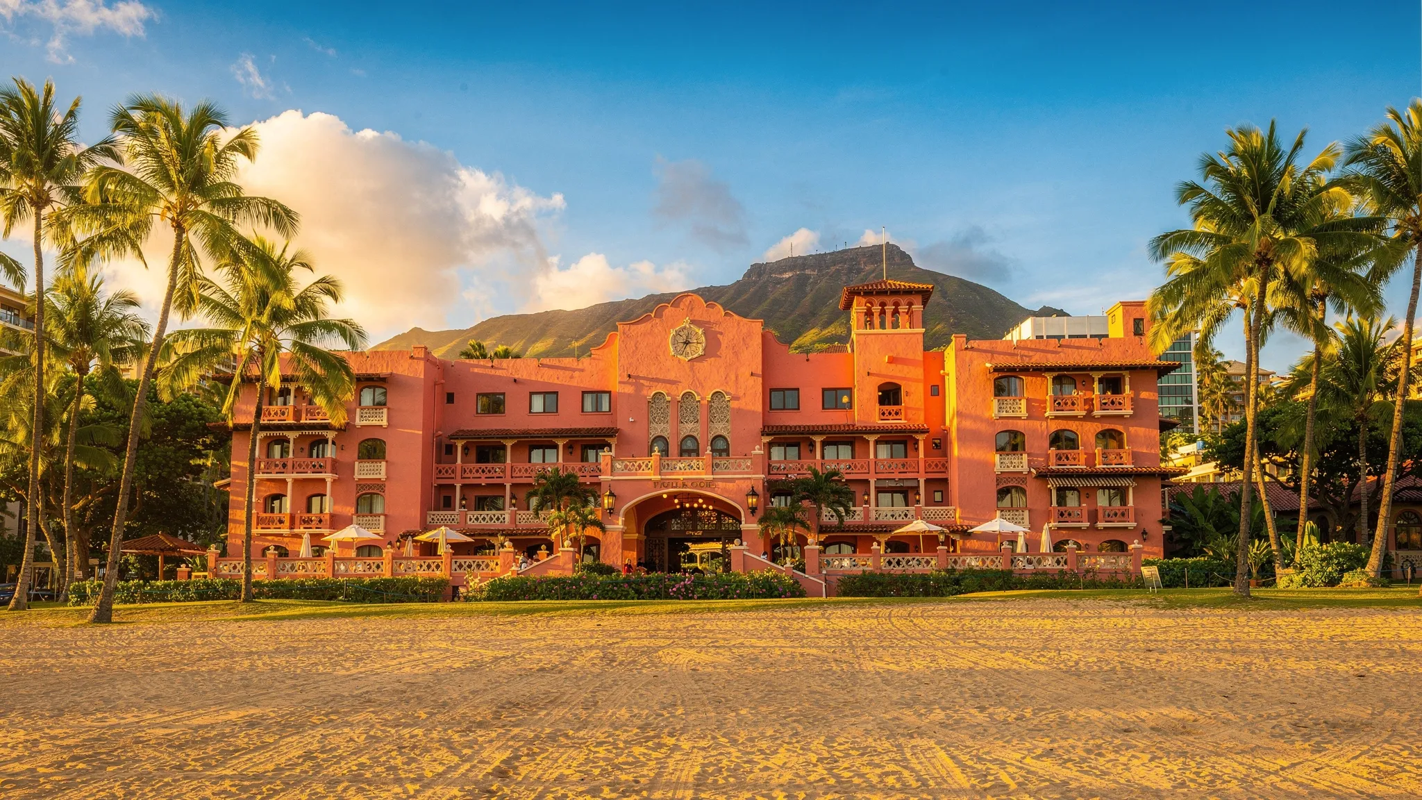The coral pink Royal Hawaiian Hotel framed by palm trees and Waikiki sand, with arched windows, shaded lanais, and Diamond Head in the distance under warm afternoon light.