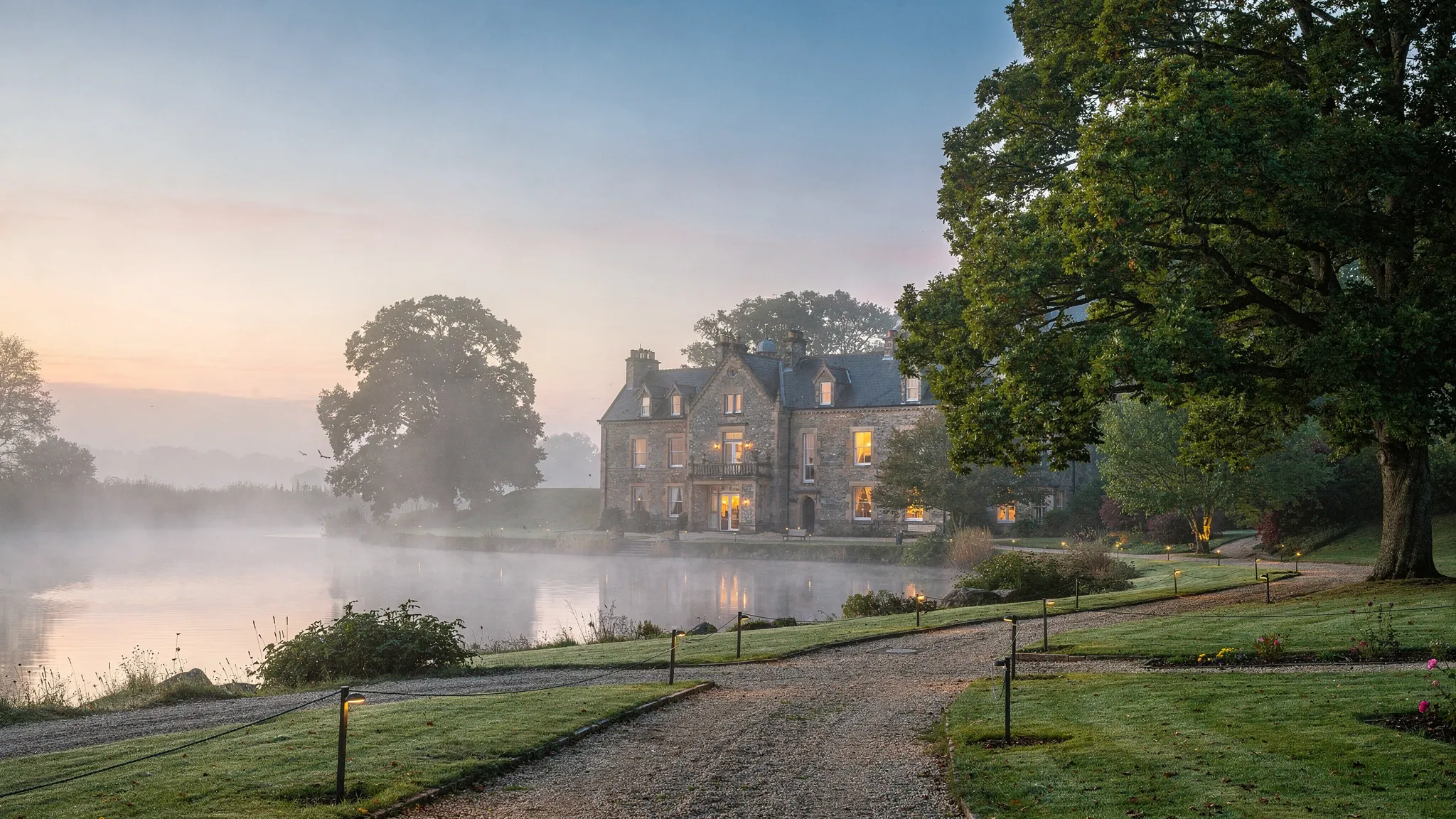A quiet country house hotel at dawn surrounded by lawns, mature trees, and a misty lake, with gravel paths leading toward a historic stone mansion and soft countryside light across the grounds.