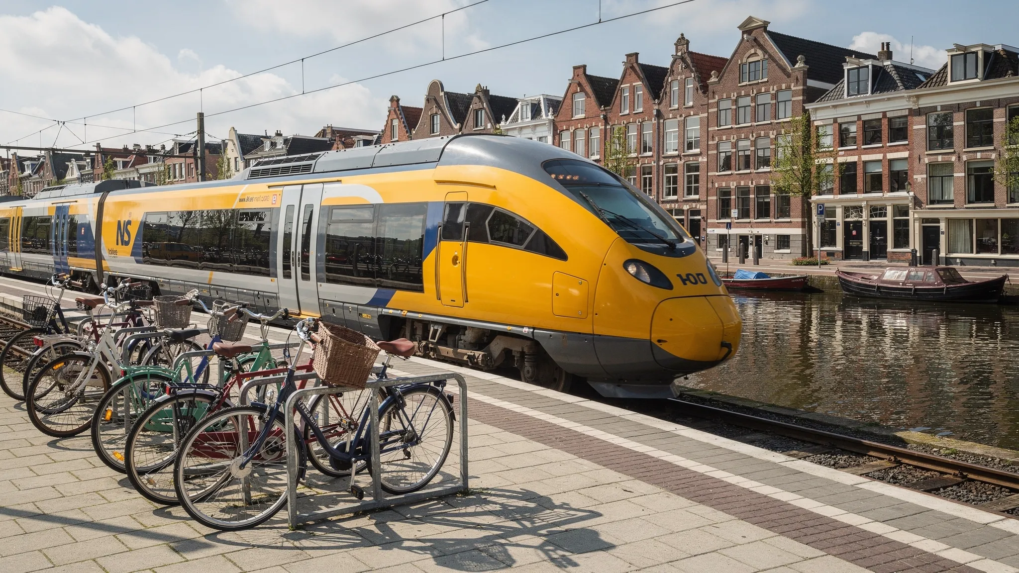 A Dutch intercity train pulling into a clean station beside bicycles, with a canal and brick buildings visible beyond the platform.