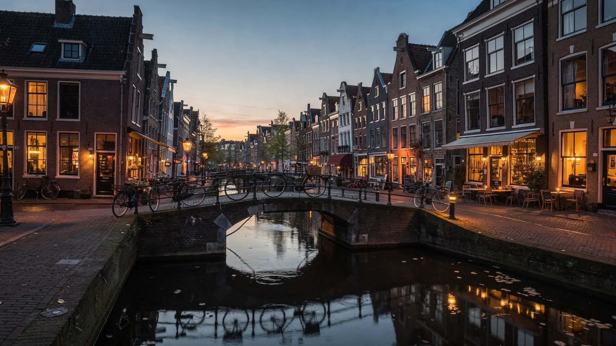 A quiet Dutch canal in an old town at evening, with bicycles on a stone bridge, warm restaurant lights, and narrow brick houses reflected in the water.