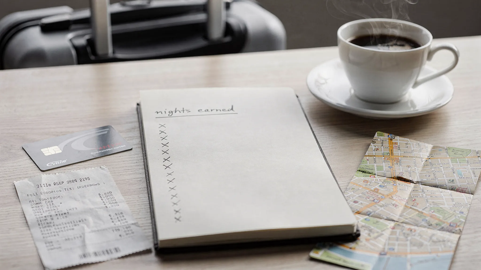 A travel-themed scene showing a business traveler’s open notebook with a simple “nights earned” tally, a hotel keycard, a receipt, and a city map on a desk next to a coffee cup and a suitcase handle in the background.