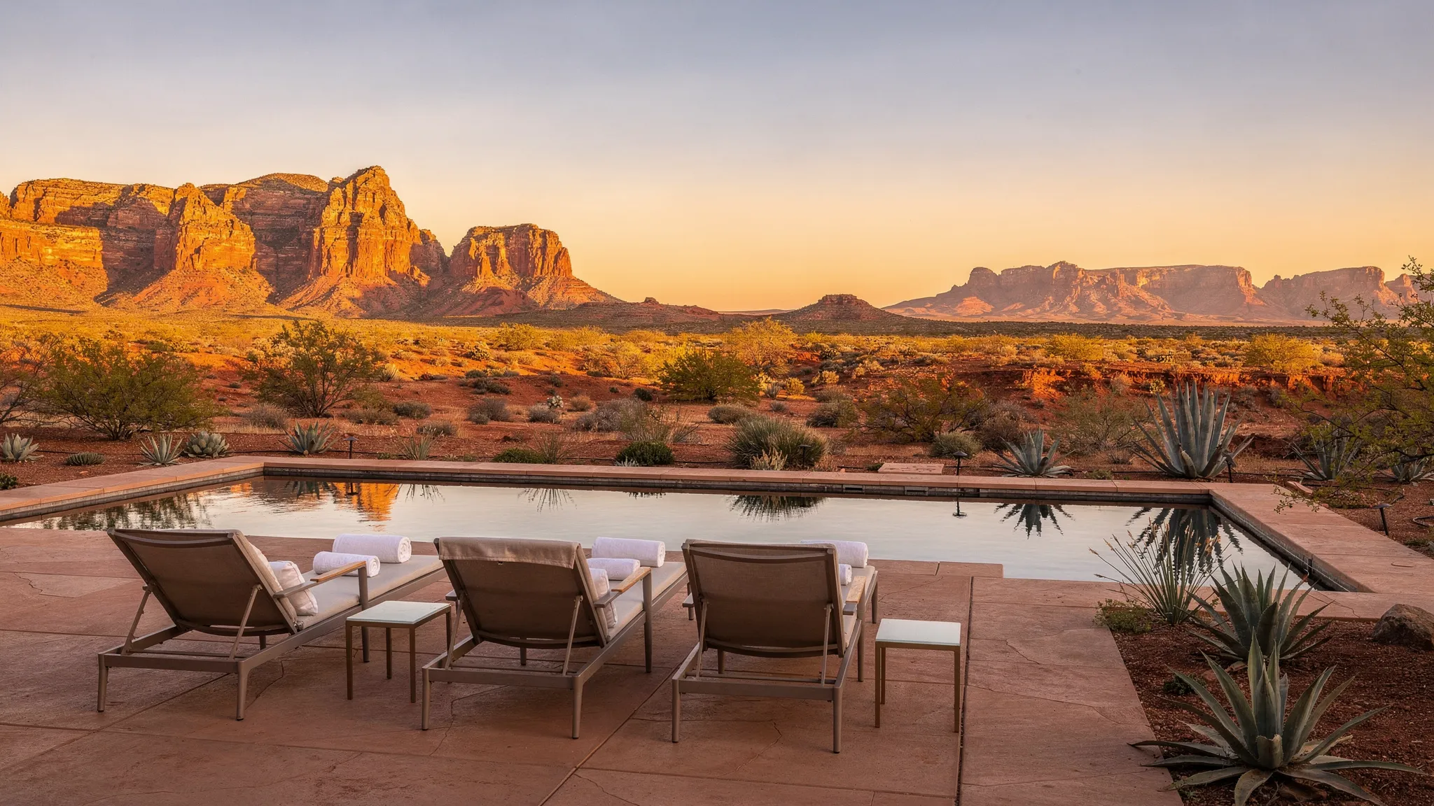 A desert wellness scene at sunrise with red rock formations in the distance, a quiet outdoor terrace with lounge chairs and a small reflecting pool, suggesting calm, warmth, and spacious air.