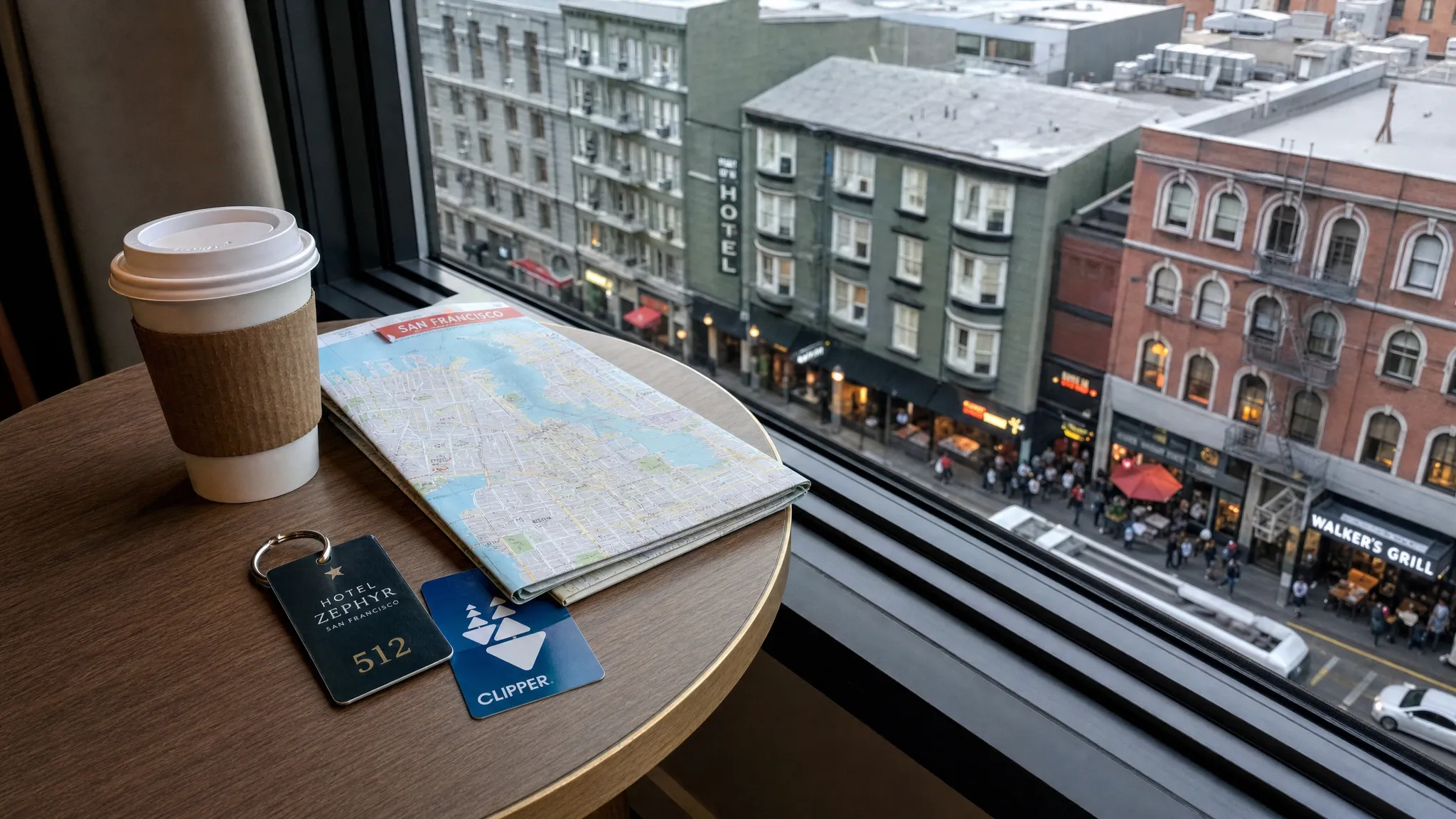 A hotel room key, folded city map, transit card, and coffee cup on a small table beside a window overlooking a dense city neighborhood with restaurants and transit lines below.