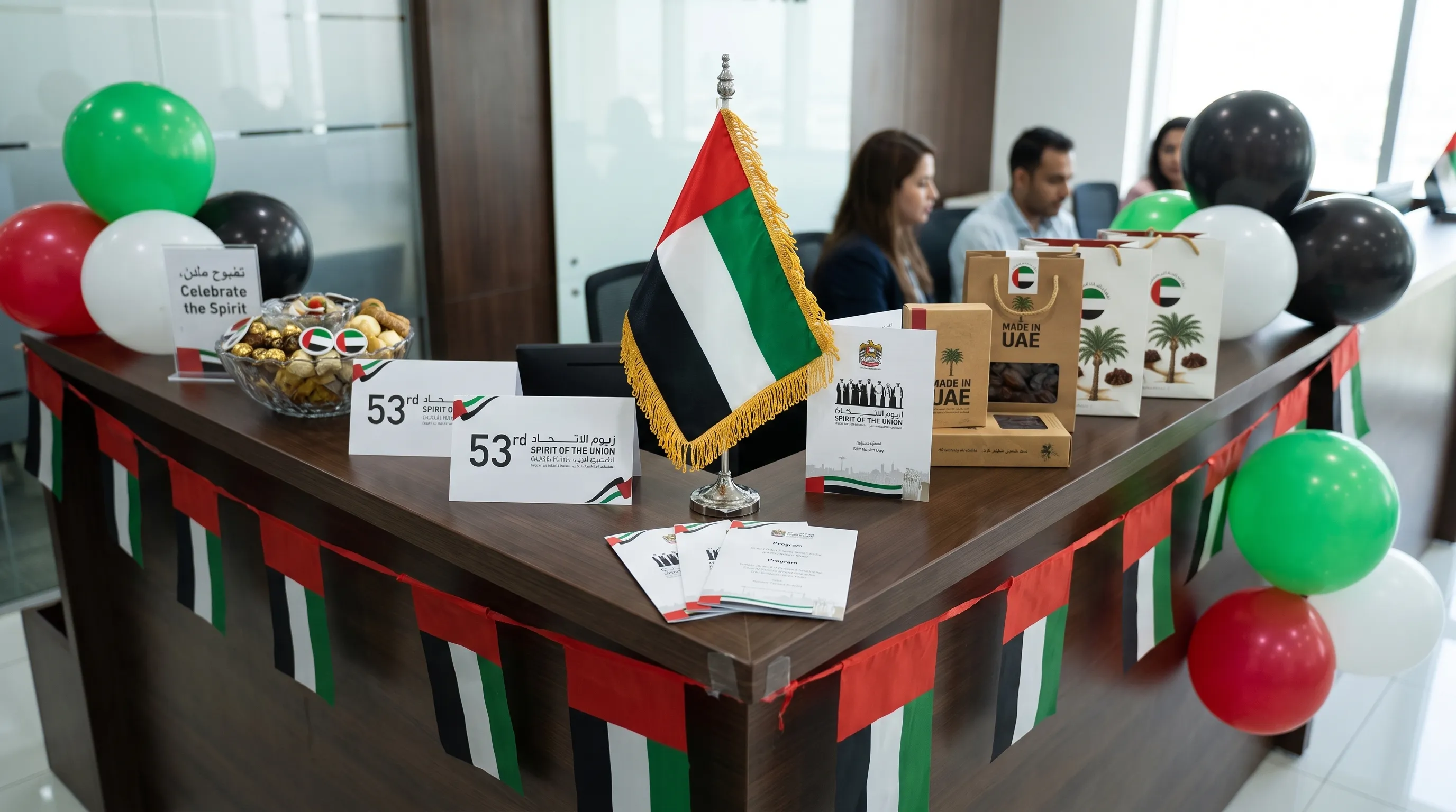 A neat UAE national occasion setup with a small UAE table flag on a reception counter, event stationery, simple festive decor and shopping items arranged for an office celebration, viewed from a slightly higher angle with the flag and sign area centered.