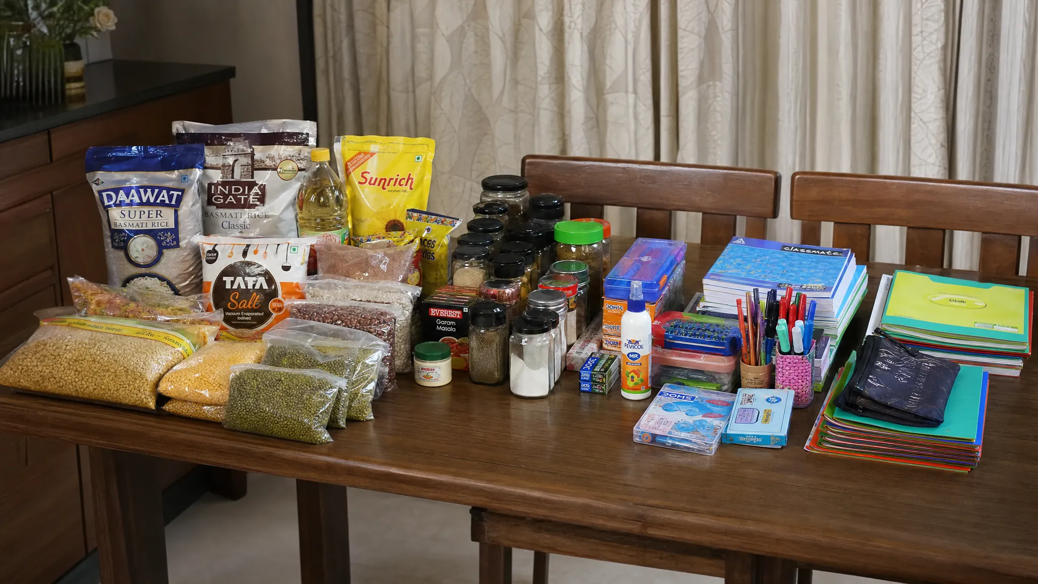 A side view of a kitchen table with groceries, rice, spices, notebooks, pens, folders, and school supplies grouped into separate sections for one weekly shopping plan.