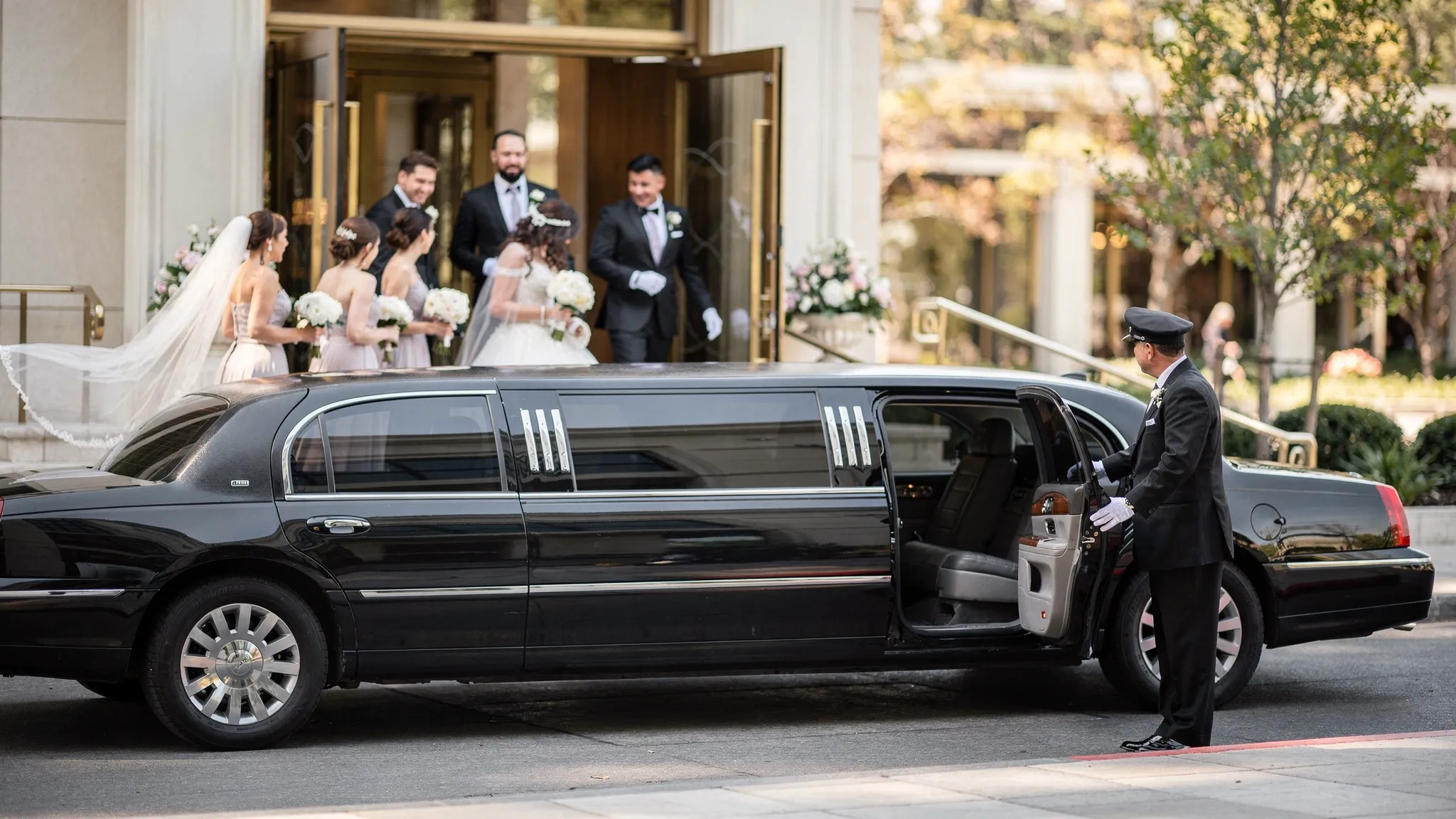A wedding party steps out of a polished black limousine outside a venue entrance, with a chauffeur opening the door and the scene focused on the vehicle and elegant arrival.