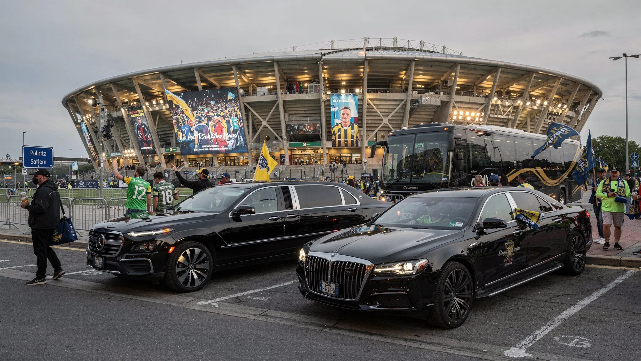 A luxury black SUV, stretch limousine, and party bus lined up near a major soccer stadium as fans in team colors arrive for an international match day.