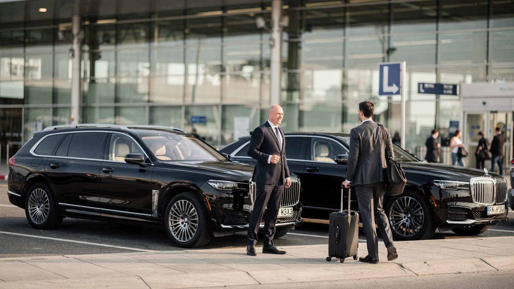 A luxury black executive SUV and sedan waiting outside a modern airport terminal while a business traveler with luggage approaches the professional chauffeur.