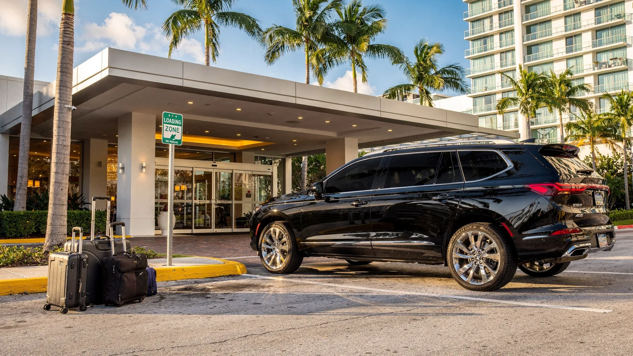 A black luxury SUV waits at a hotel porte cochere in Miami with clear loading-zone signage, luggage beside the curb, and palm trees and modern buildings in the background.