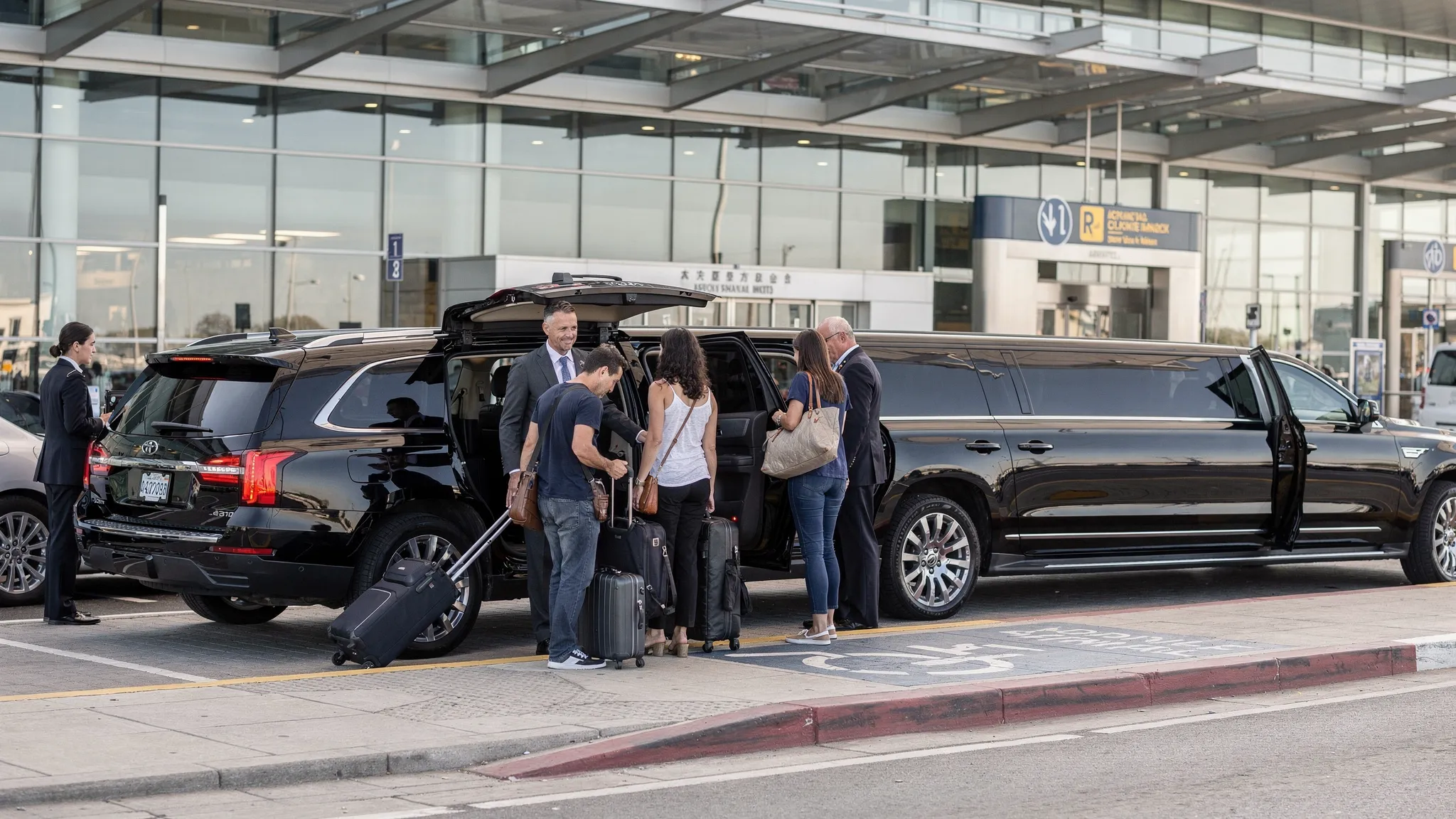 A luxury black SUV and stretch limousine waiting at an airport arrivals curb with travelers loading luggage into the vehicles.