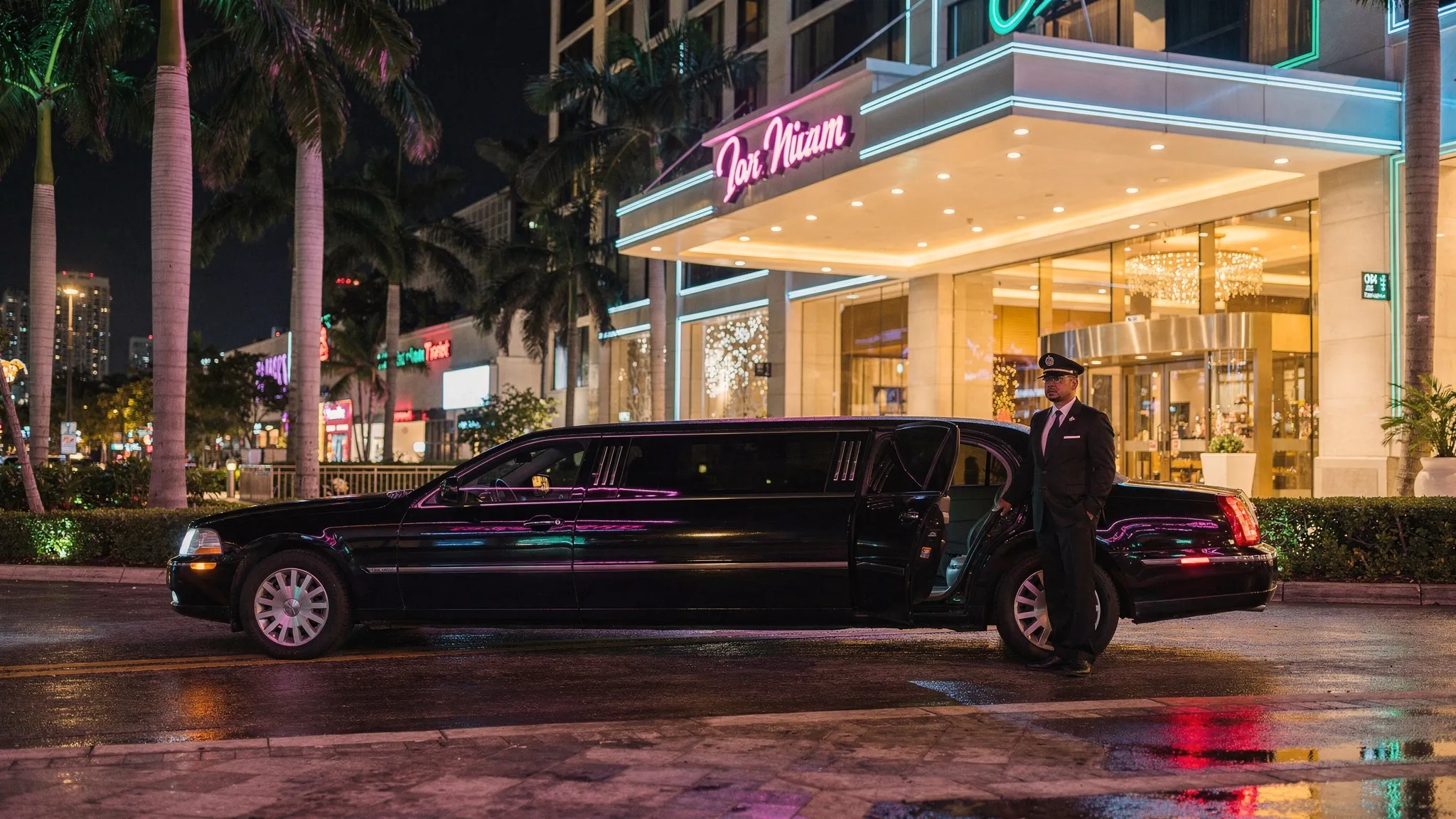 A glossy black stretch limousine pulled up at a Miami hotel porte-cochere at night, with palm trees, neon reflections, and a chauffeur standing by the rear passenger door.
