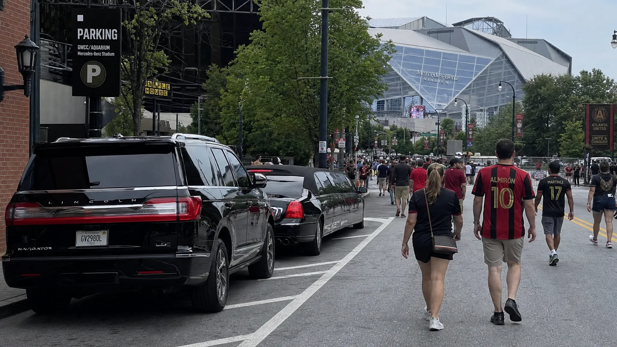 A black luxury SUV and a stretch limousine parked along a downtown Atlanta street with Mercedes-Benz Stadium visible in the distance, while adult fans walk toward the venue on a busy soccer matchday.