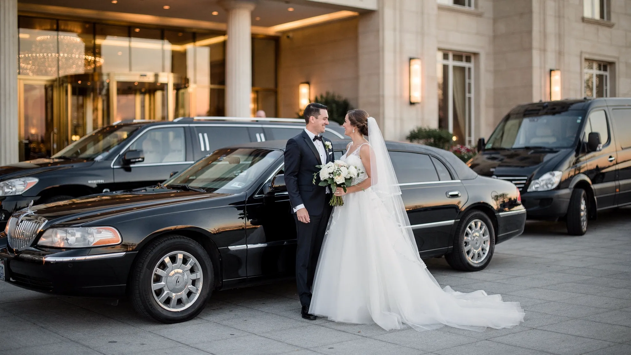 A newly married couple standing beside an elegant black limousine outside a wedding venue, with a luxury SUV and Sprinter van parked nearby for the wedding party and guests.