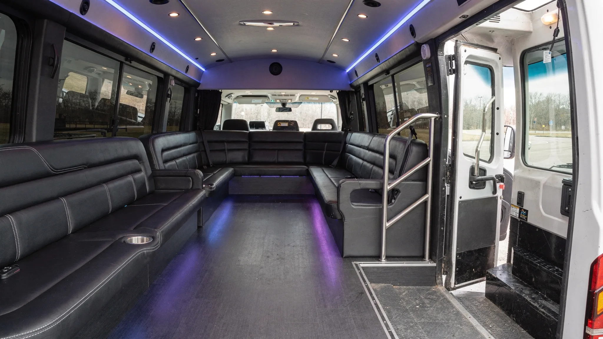 A clean, modern party bus interior with perimeter seating, LED accent lighting, cupholders, and clear aisle space, parked with the door open to show the entry steps and safety handrails.